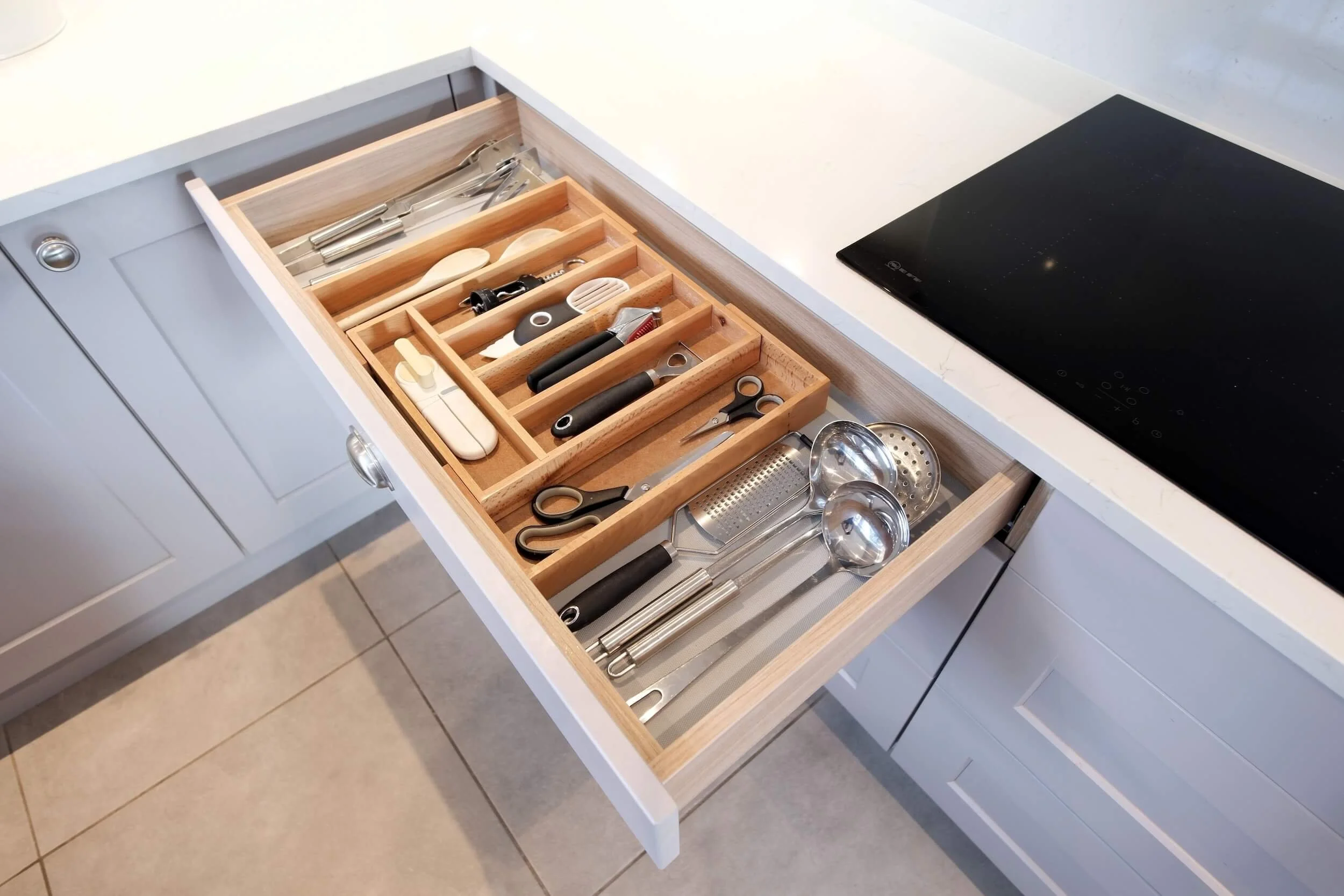 Kitchen drawer open to reveal organised utensils and tools in wooden dividers, including scissors, ladles, and measuring spoons.