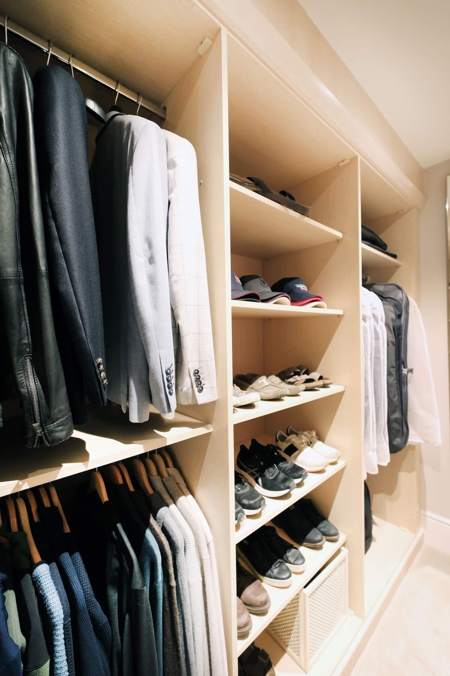 Clothes hanging neatly in a mens closet in a walk in dressing room and shoes and hats arranged neatly on shelves. 