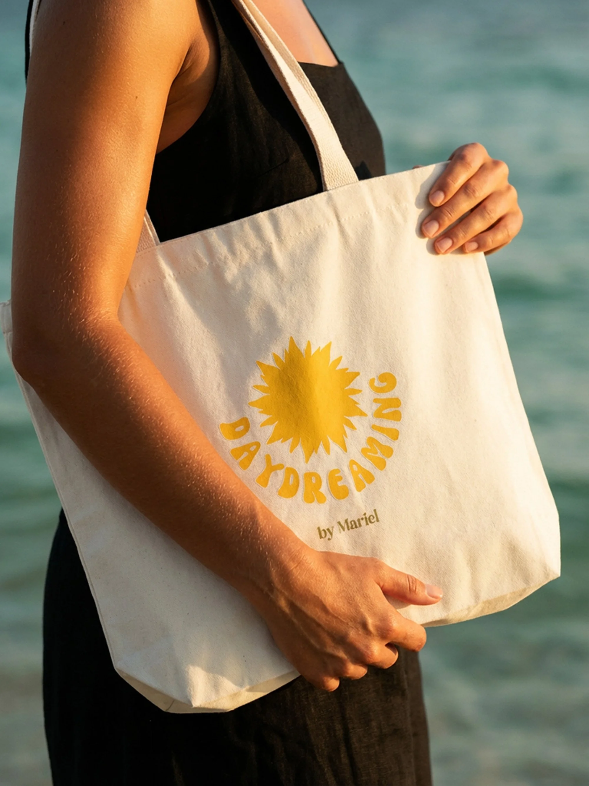 A person holding a tote bag with a yellow sun design and the text 'Daydreaming' and 'by Mariel' printed on it, standing by the water.