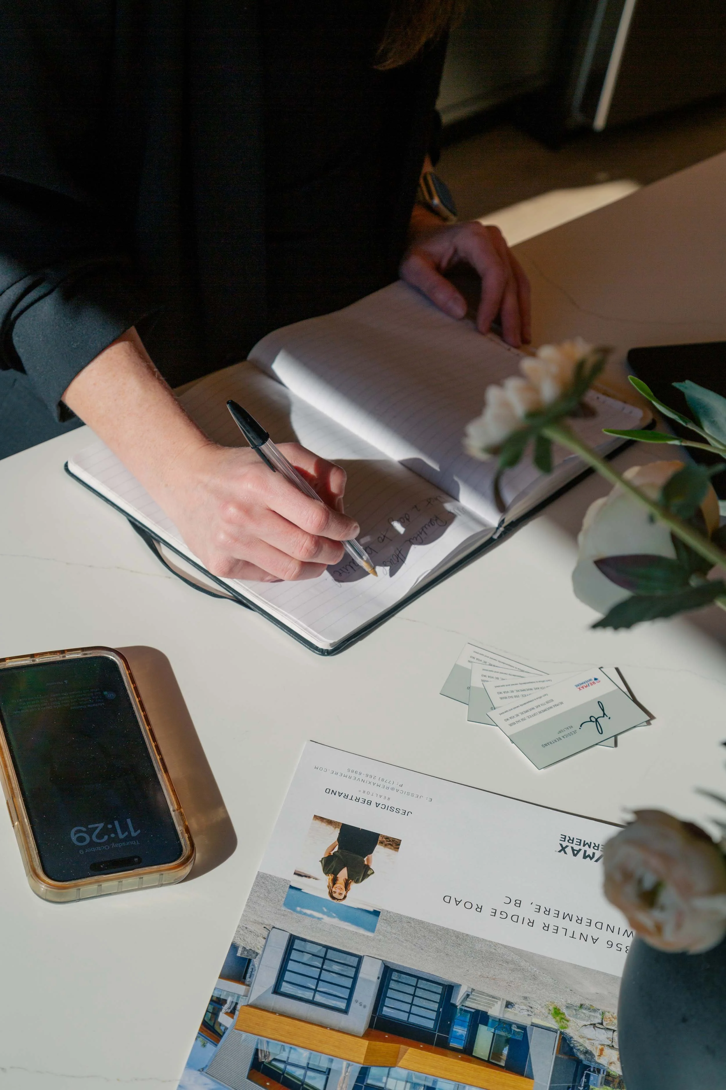Flatlay of Jessica Bertrand’s RE/MAX Invermere real estate materials, including her brochure with photo, a stack of business cards, a smartphone, and an open notebook on a white desk.