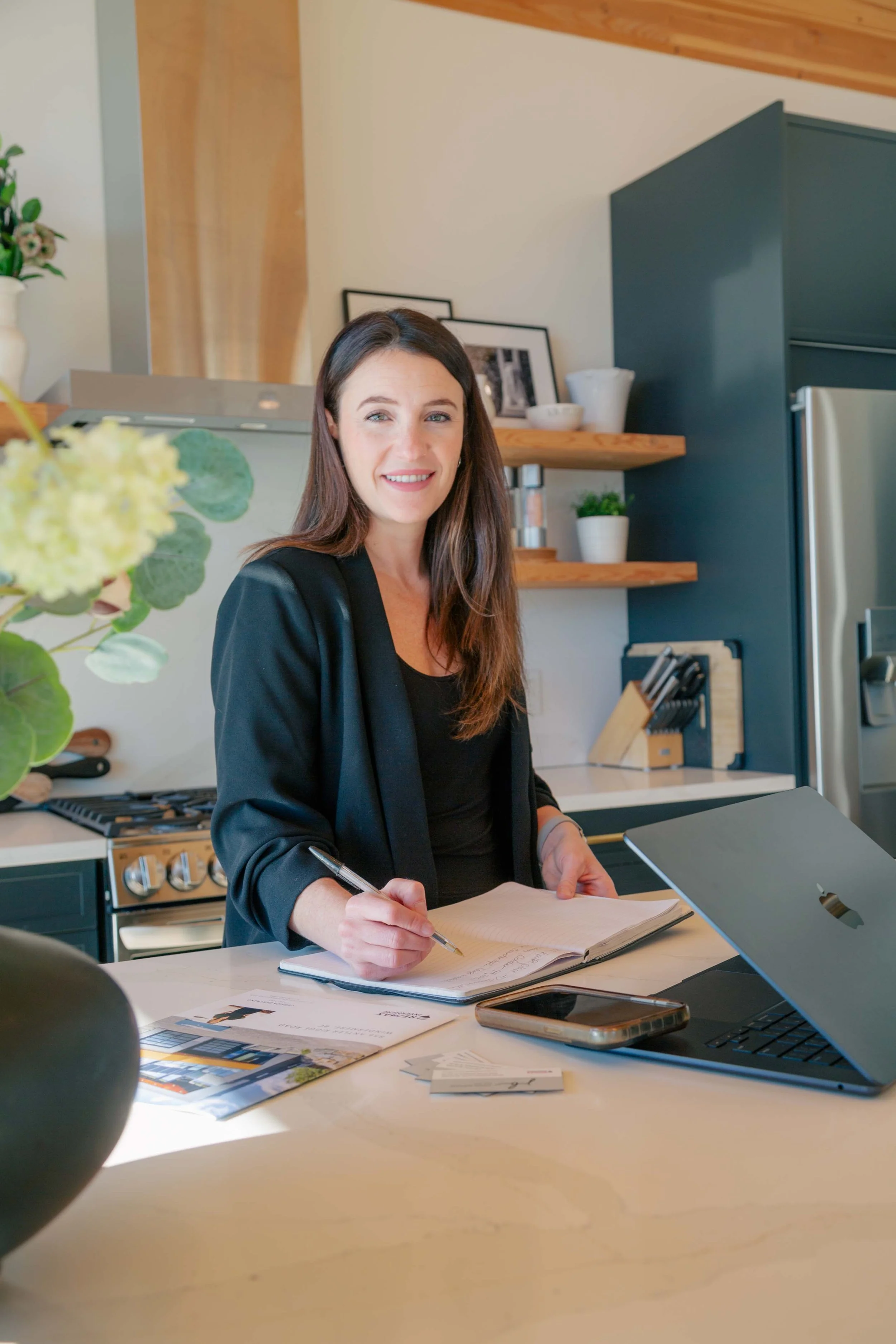 Jessica Bertrand, a RE/MAX Invermere real estate agent, smiling while writing in a notebook in a modern kitchen. She is wearing a black blazer and standing beside open shelves and stainless-steel appliances, with her laptop open nearby.