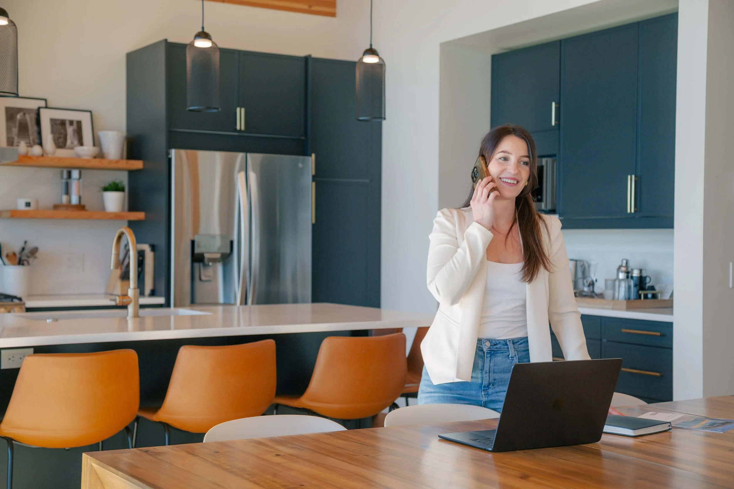 Jessica Bertrand, a RE/MAX Invermere real estate expert, standing in a modern kitchen while speaking on the phone. She is smiling, wearing a light blazer and jeans, with an open laptop on the counter in front of her.