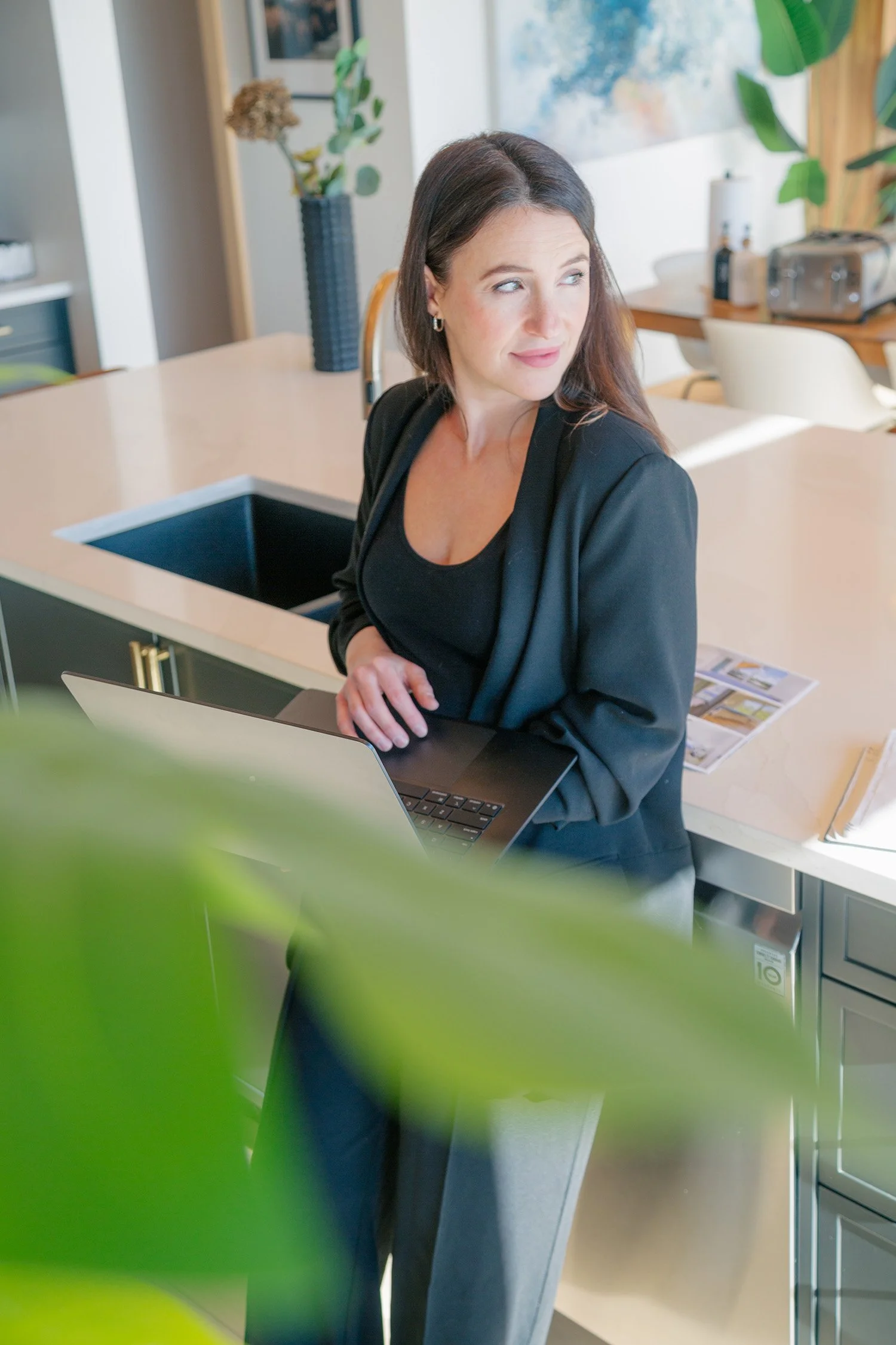 Jessica Bertrand, a RE/MAX agent serving the Invermere BC real estate market, standing in a modern kitchen with a laptop in hand. She is wearing a black blazer, looking thoughtfully to the side, with soft greenery in the foreground.