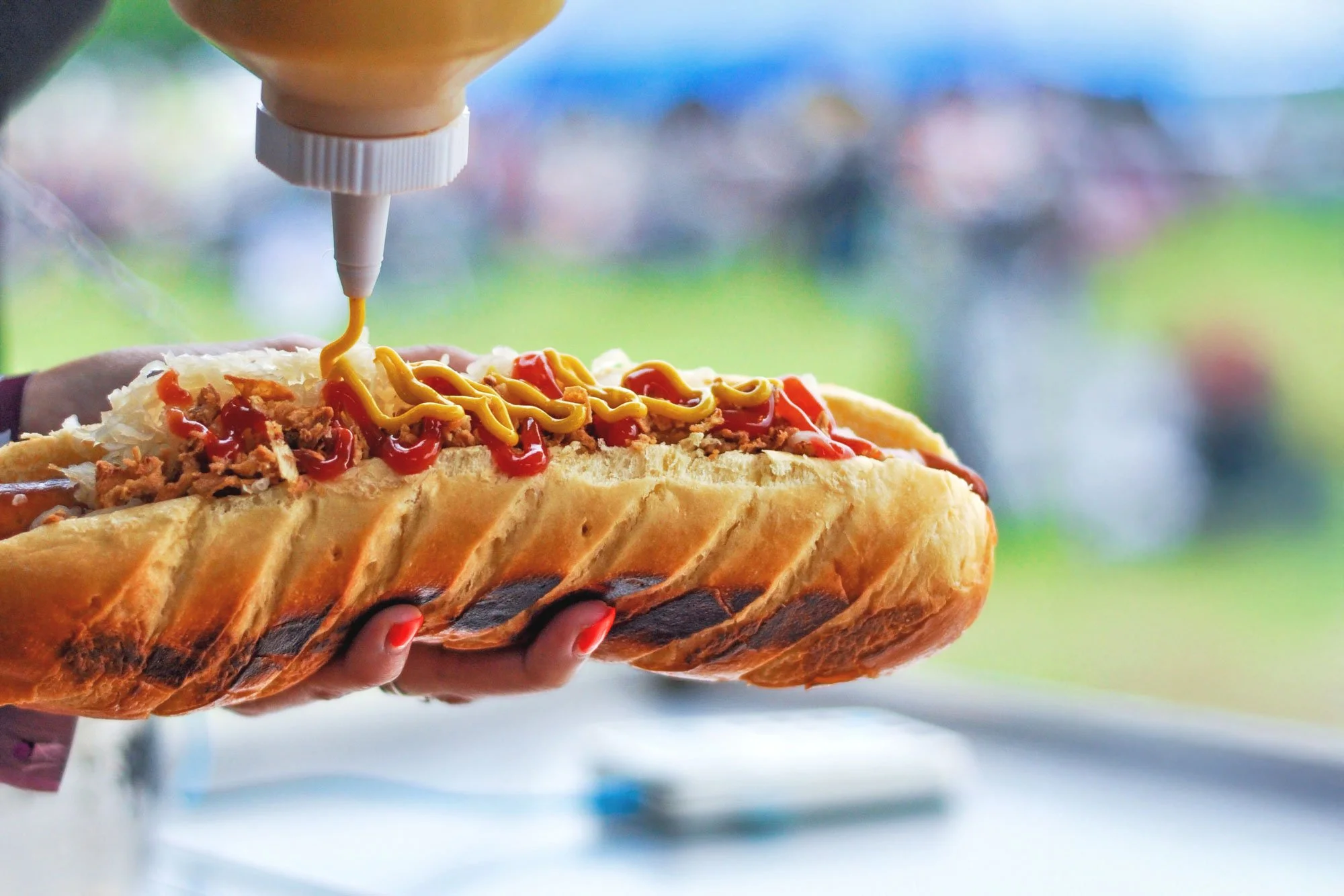 close up of mustard being added to a hot dog with a blurred festival scene in the background