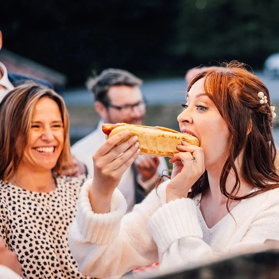 Bride taking a bite of a hot dog during the evening wedding reception while guests smile in the background