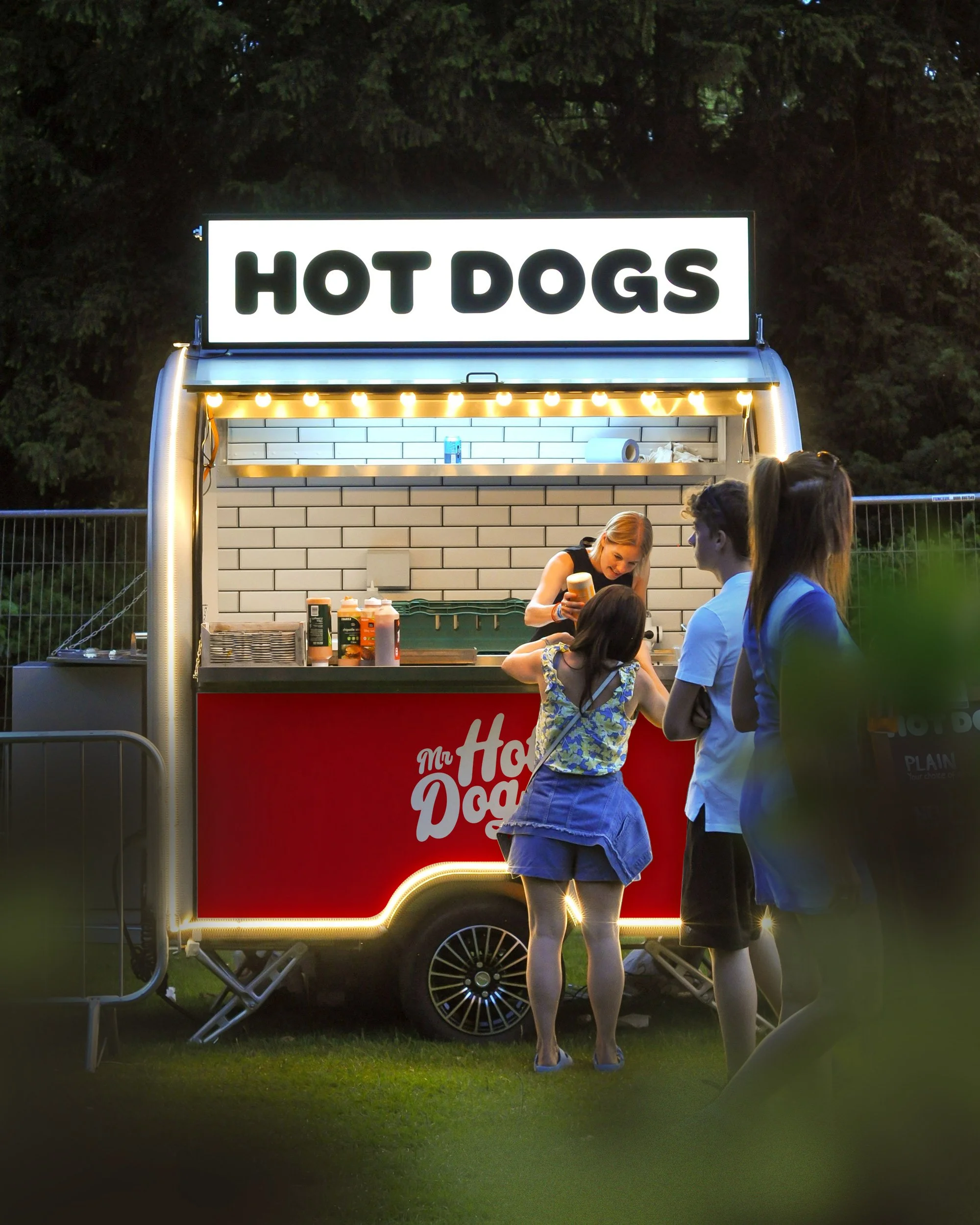 A hot dog food stand with a large illuminated sign reading 'HOT DOGS'. There are two people standing at the stand, with a woman preparing food inside the stand.
