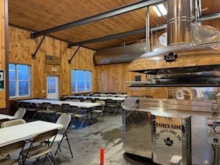 Interior of a sugarhouse with wooden walls, multiple windows, and several tables and chairs arranged for pancake breakfast.