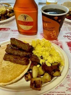 A breakfast plate with scrambled eggs, sausage patties, hash browns, and toast. In the background, there is a cup of black coffee, a bottle of syrup, and a glass of orange juice.