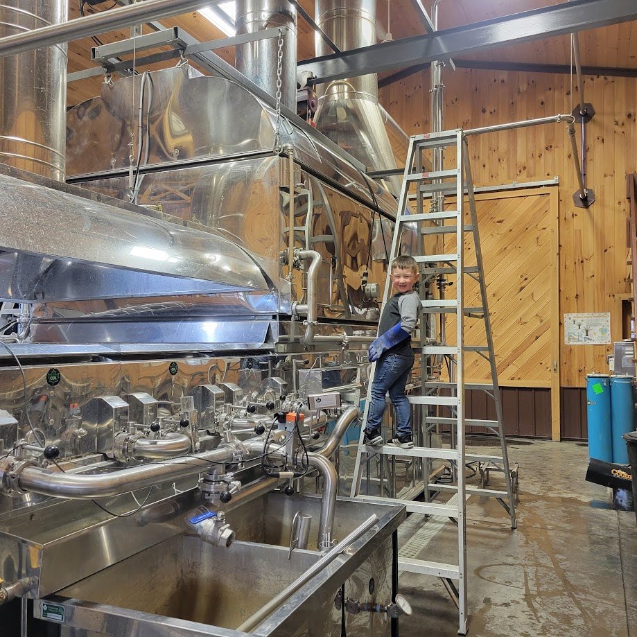 A young boy standing on a ladder inside sugarhouse, smiling at the camera, with large maple evaporator around him.