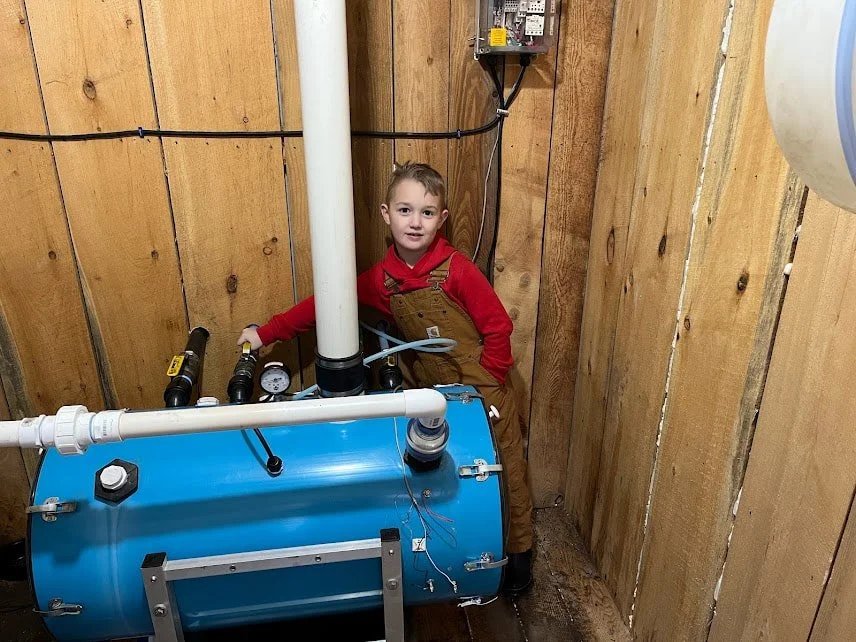 A young child in Carhartt overalls and a red shirt standing next to a large vacuum pump in a pump house collecting maple sap.