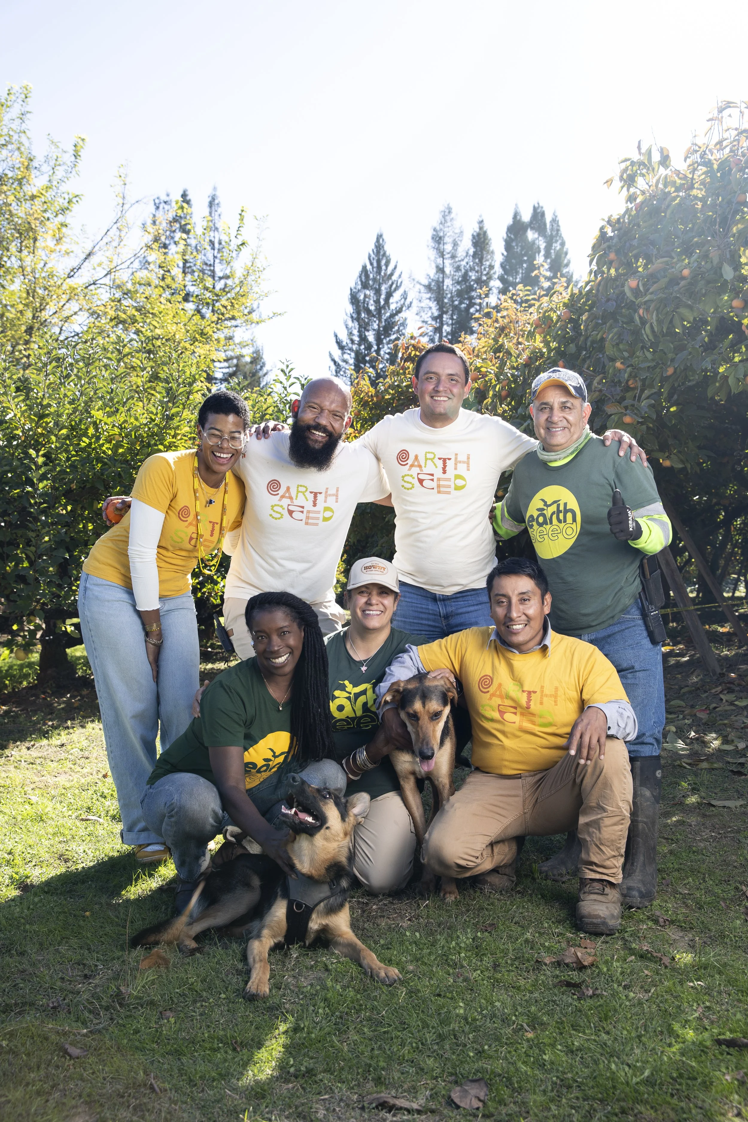 7 people, 4 standing in the back and 3 kneeling on the group wearing yellow, white or green Earthseed t-shirts with two dogs in the front