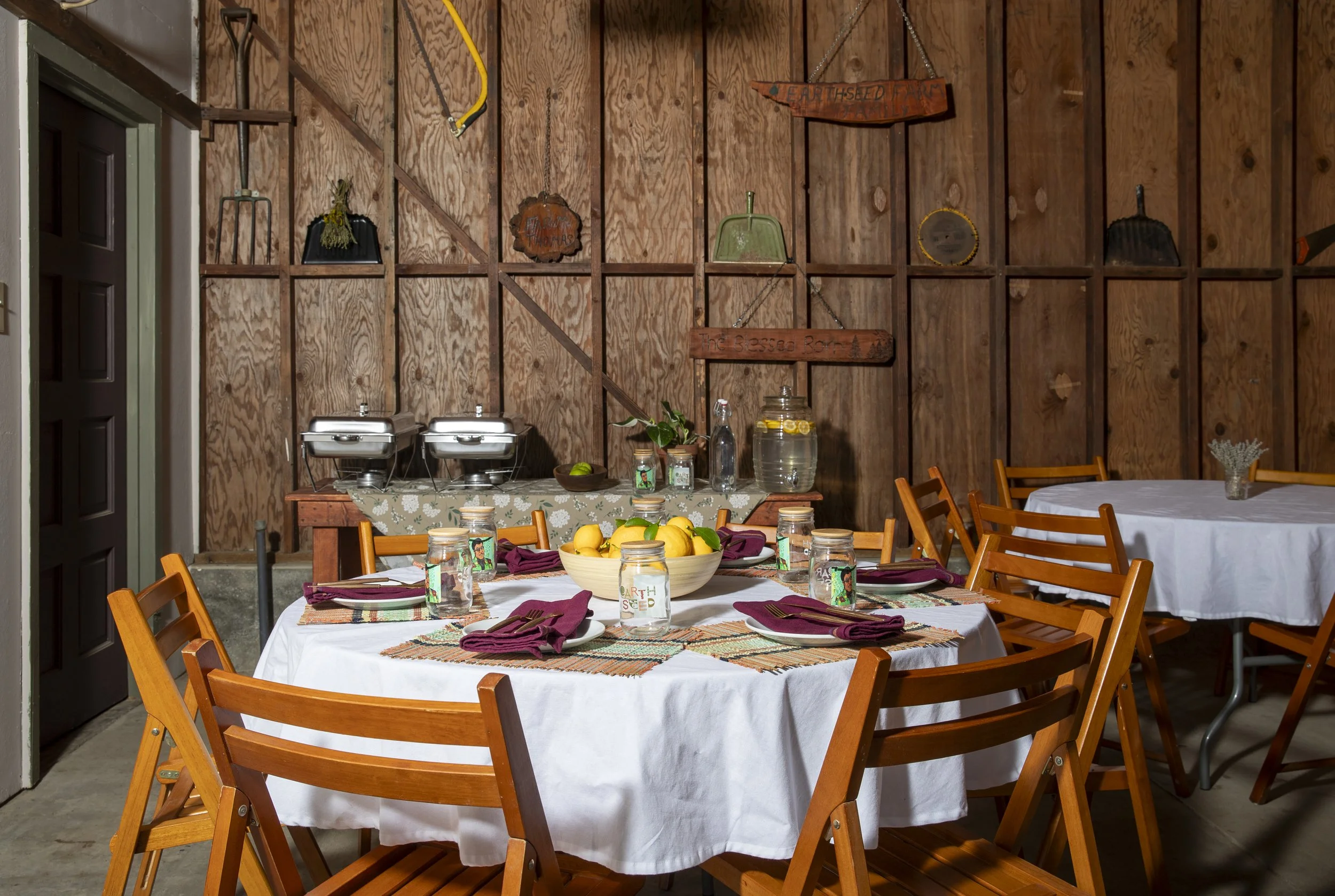 A circular table covered with a white cloth surrounded by 6 wooden chairs in front of a wood paneled wall displaying various tools. A bowl of fresh lemons, straw placemats, white small plates and maroon cloth napkins and branded jars on the table