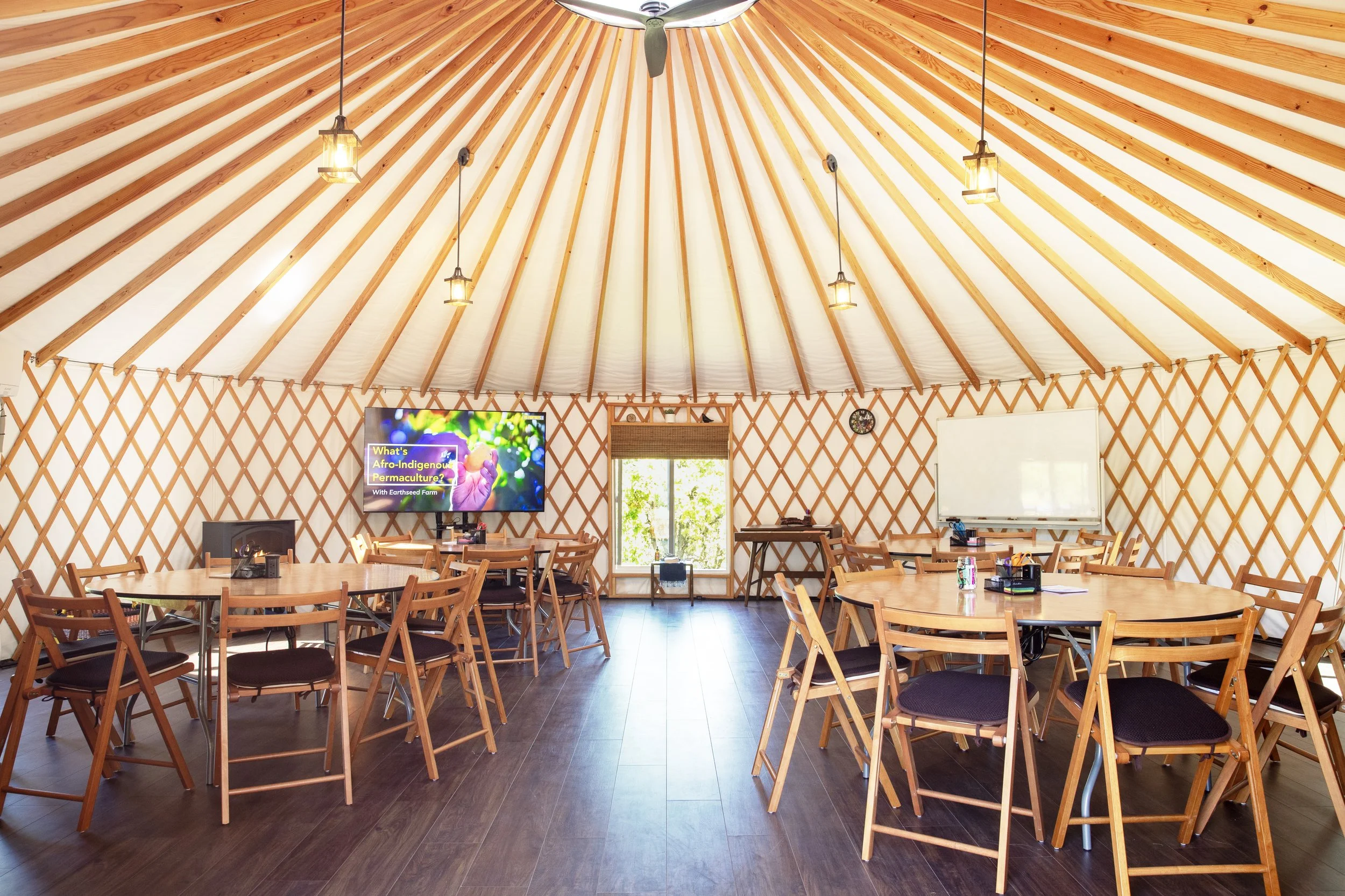 Inside 30ft yurt. Very bright natural light and wood beam coned ceiling. 4 wood circular tables surrounded by wood chairs on a dark wood floor. Big screen TV in left back side with a large window to the right of it