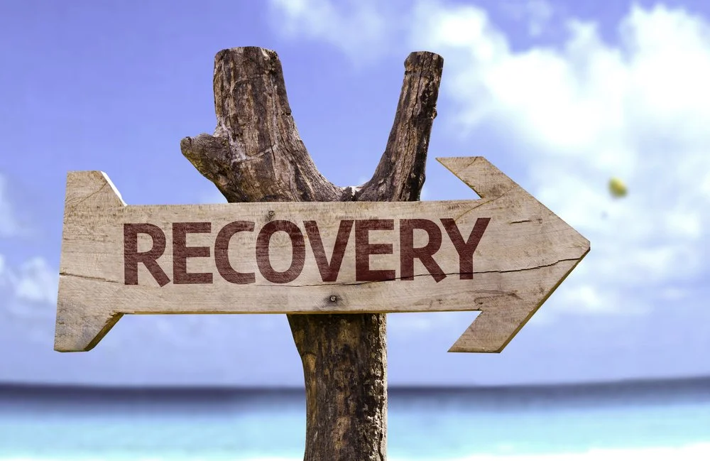 Wooden sign with the word 'RECOVERY' pointing to the right, mounted on a weathered tree branch against a beach and ocean background with a cloudy sky.