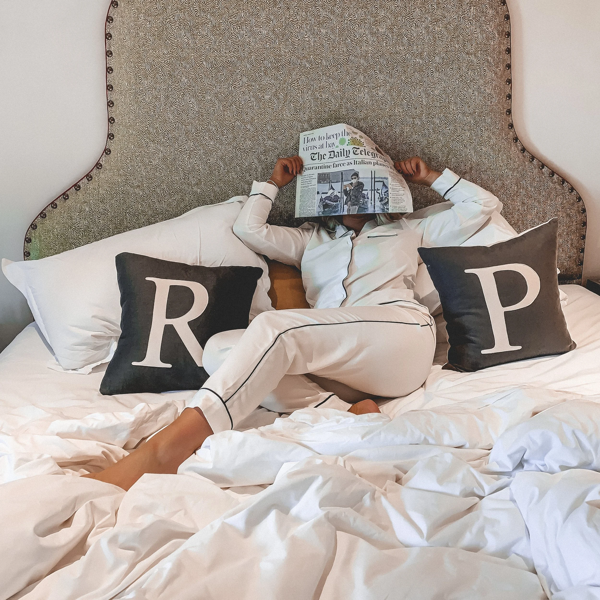 Person in white pajamas sitting on a bed with a headboard, reading a newspaper covering their face, surrounded by pillows with letters R and P, in a cozy bedroom.