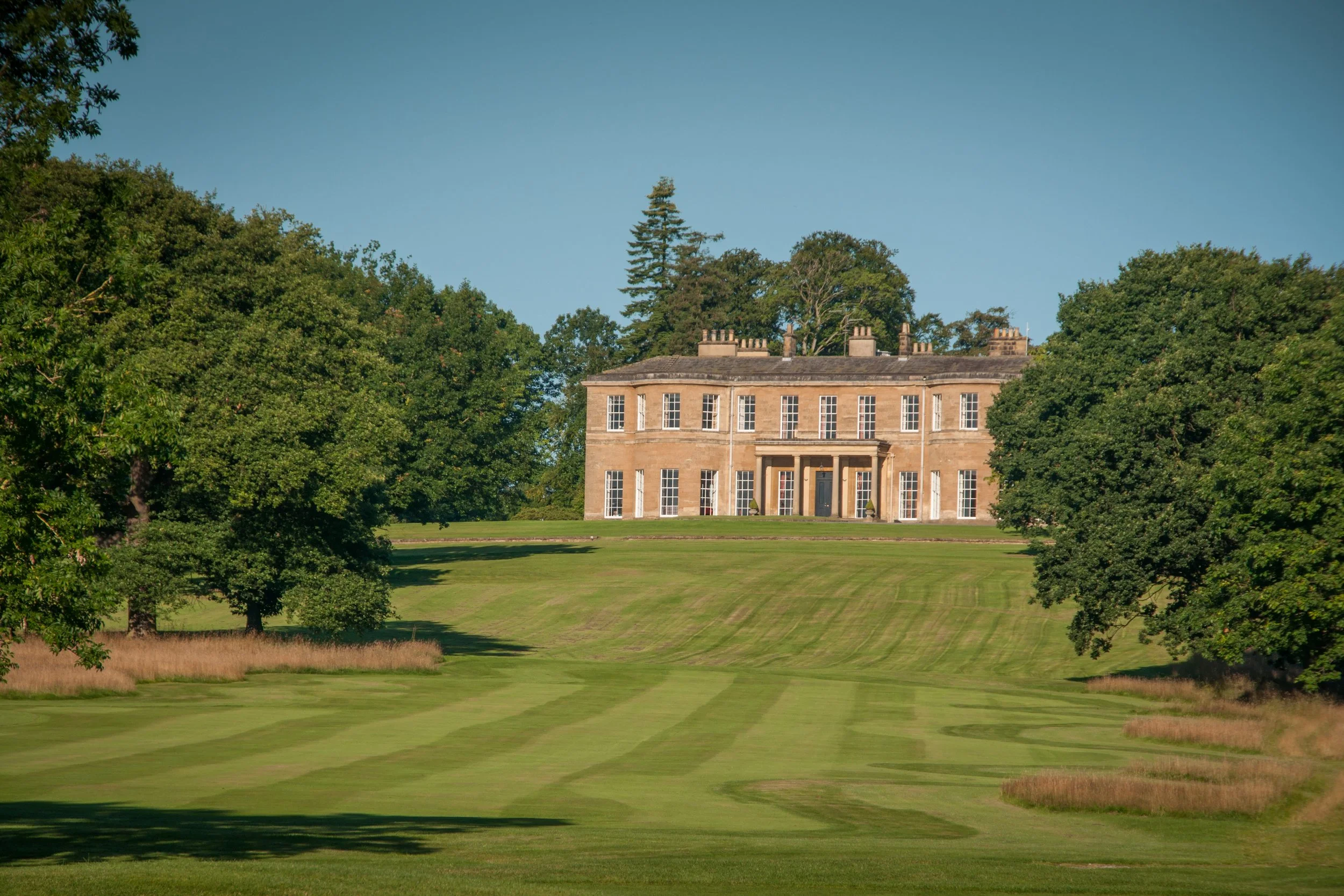 A large, historic mansion with a symmetrical facade, surrounded by tall trees and manicured lawns on a sunny day.
