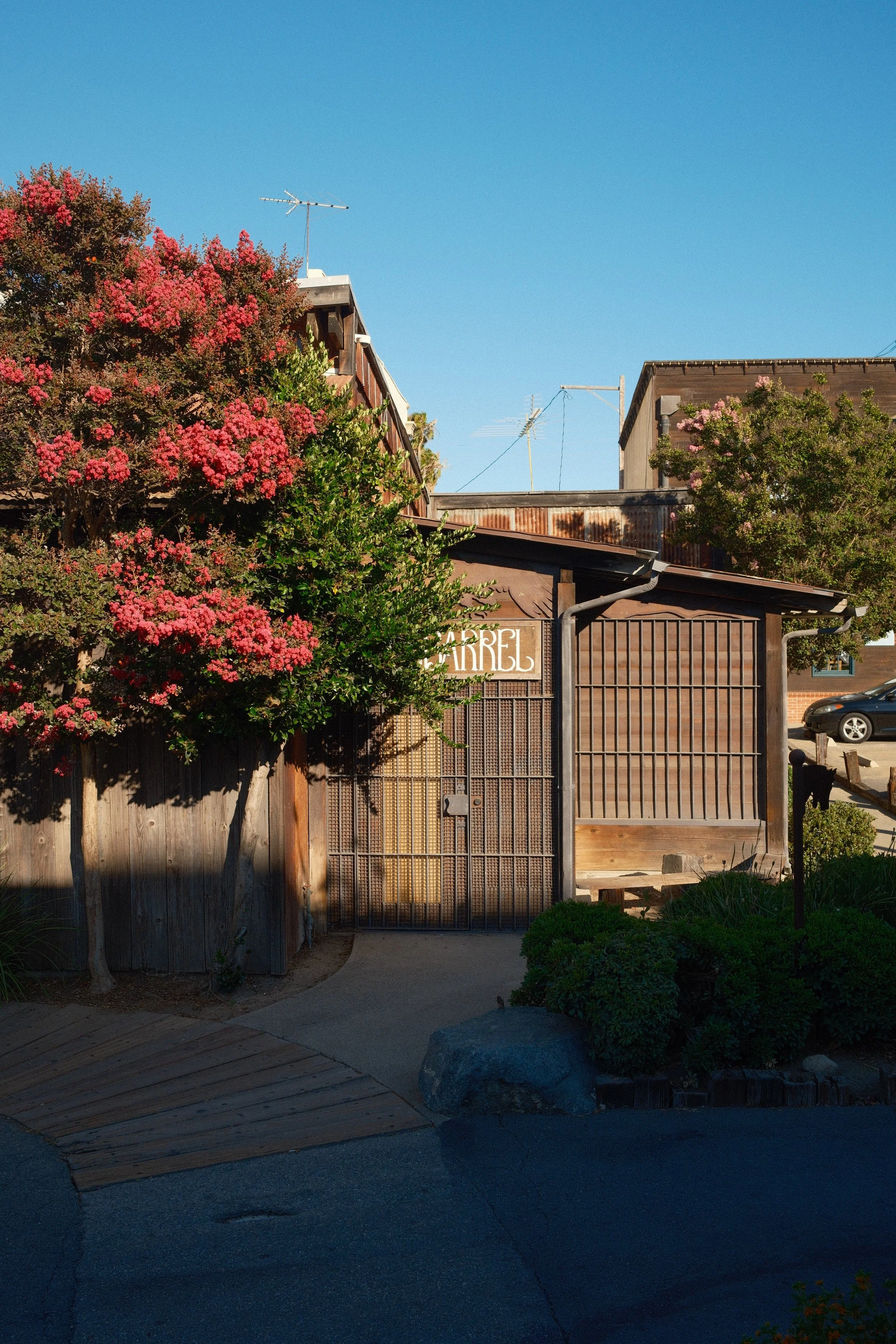 A wooden gate in front of a building with trees and pink flowers, a blue sky overhead, and a car parked on the street to the right.