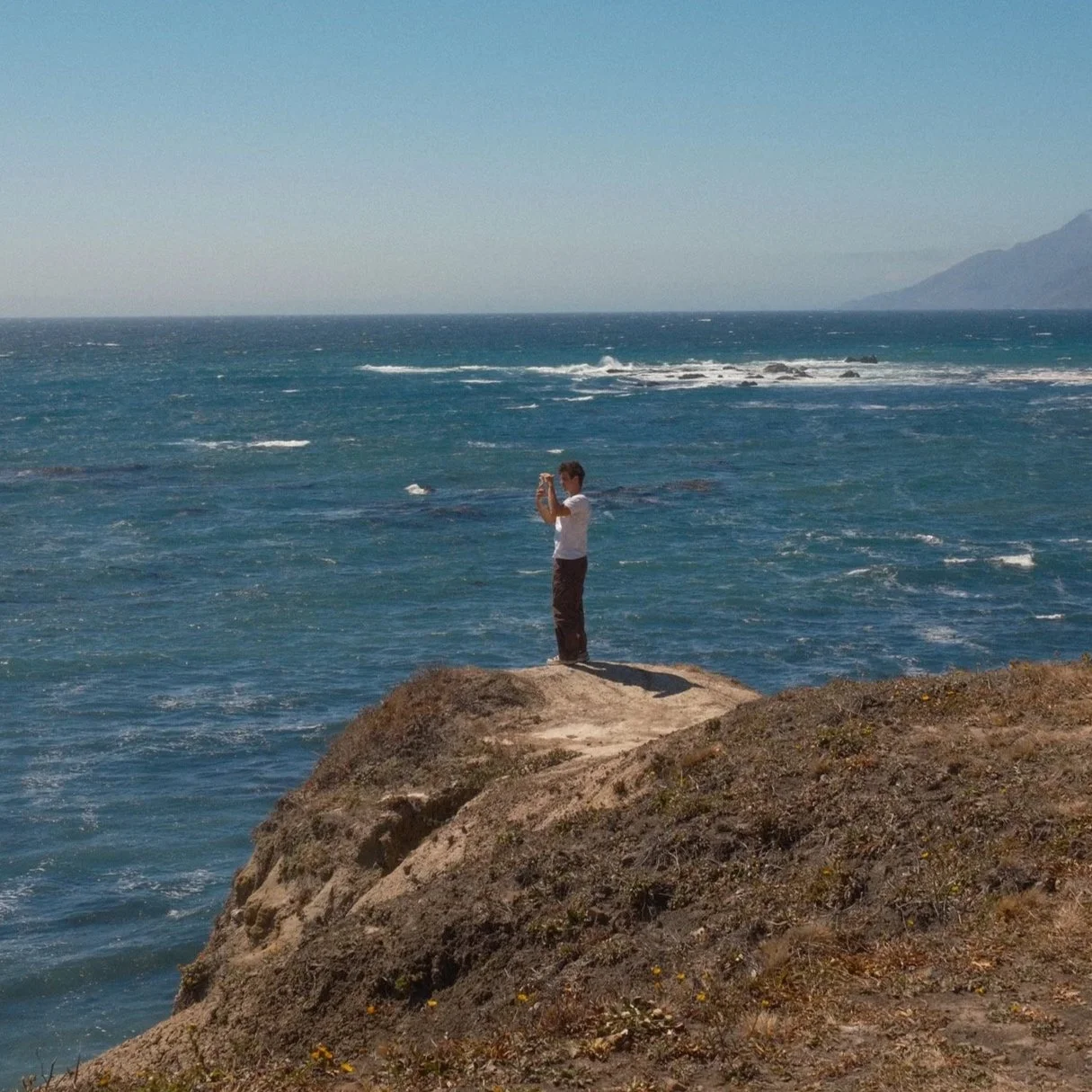 A person standing on a rocky cliff taking a photo of the ocean with mountains in the background.