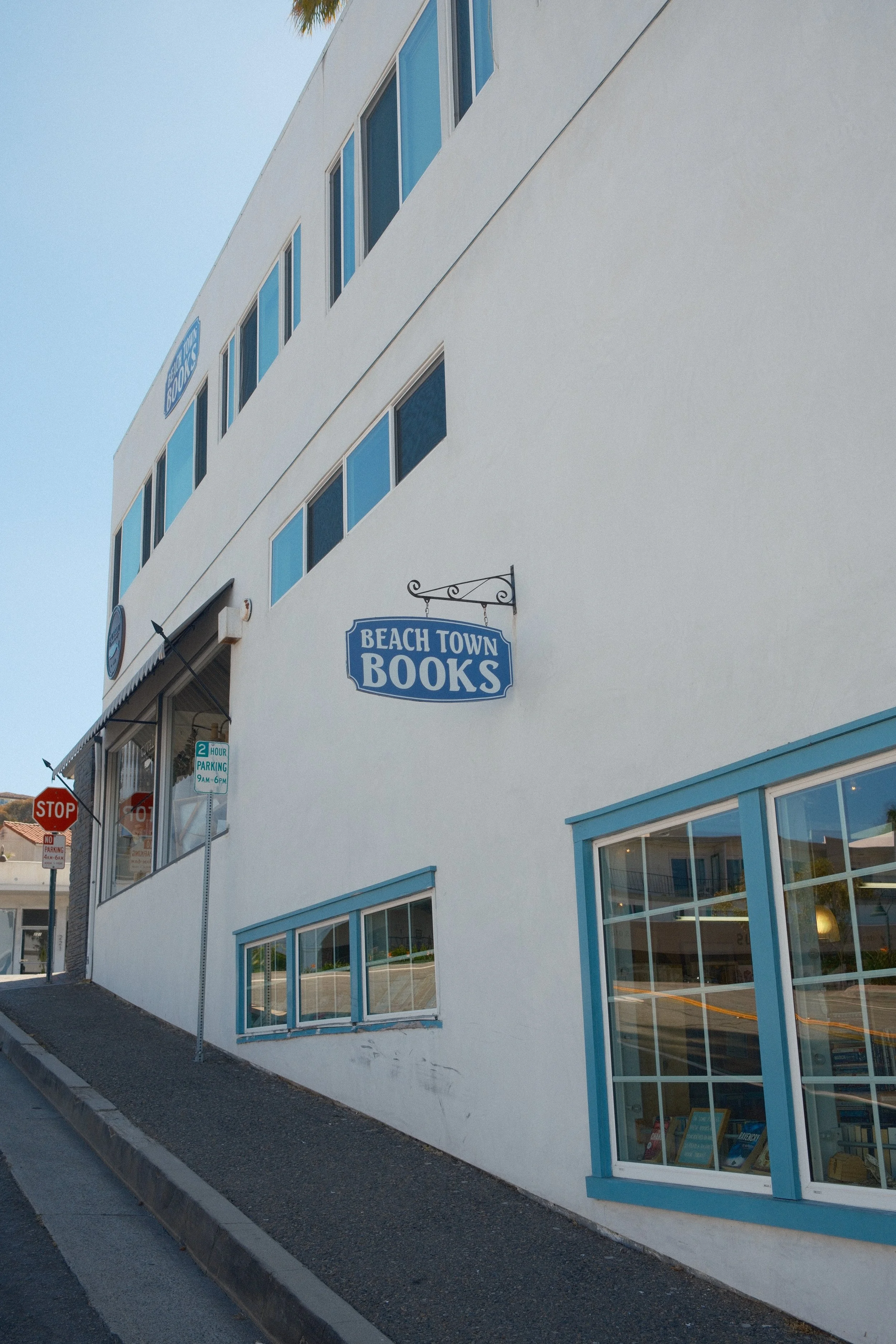 Exterior of a bookstore called 'Beach Town Books' with white walls and large glass windows, blue trim, and signs indicating parking and stopping signs nearby.