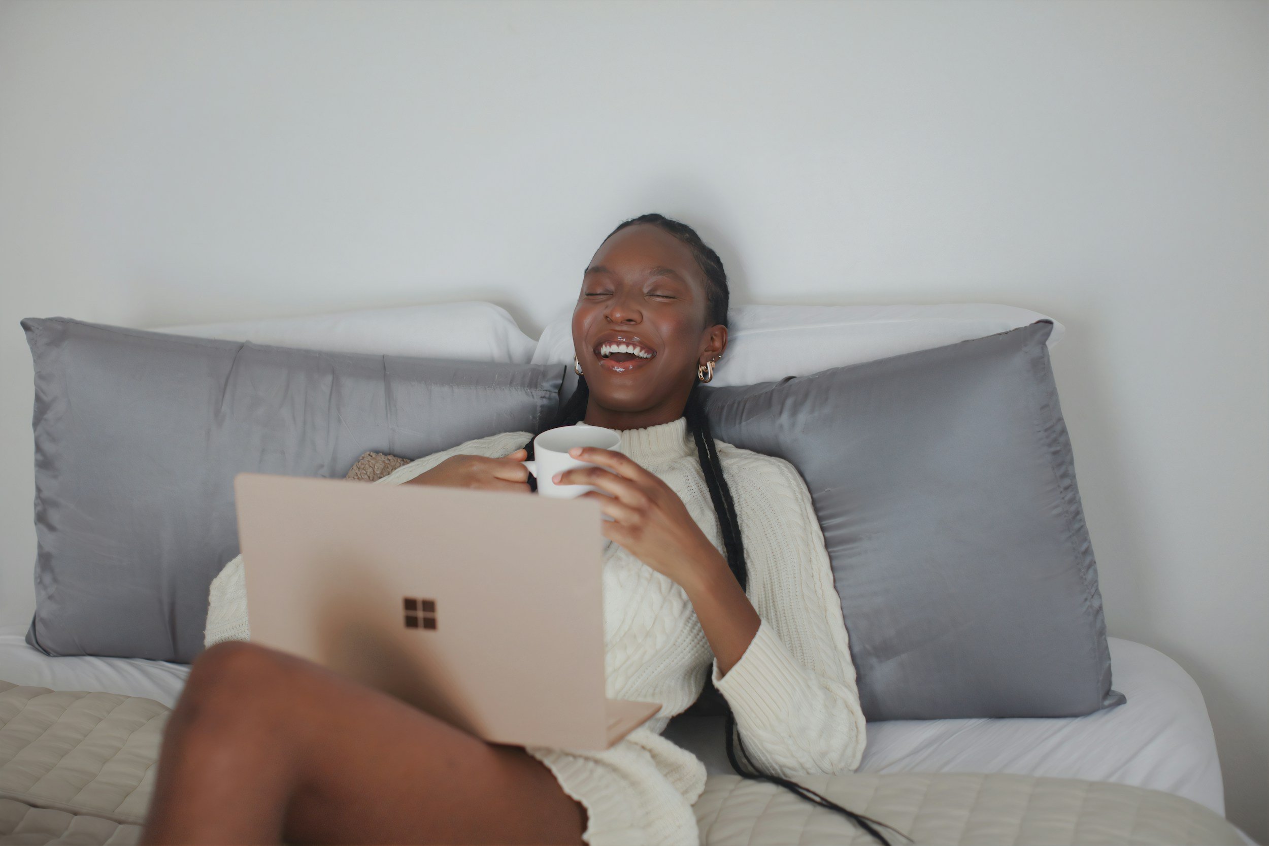 A woman with braided hair, wearing gold hoop earrings and a cream-colored sweater, is sitting on a bed, laughing, while holding a white mug. She has a laptop in front of her, and there are gray pillows behind her against a plain wall.