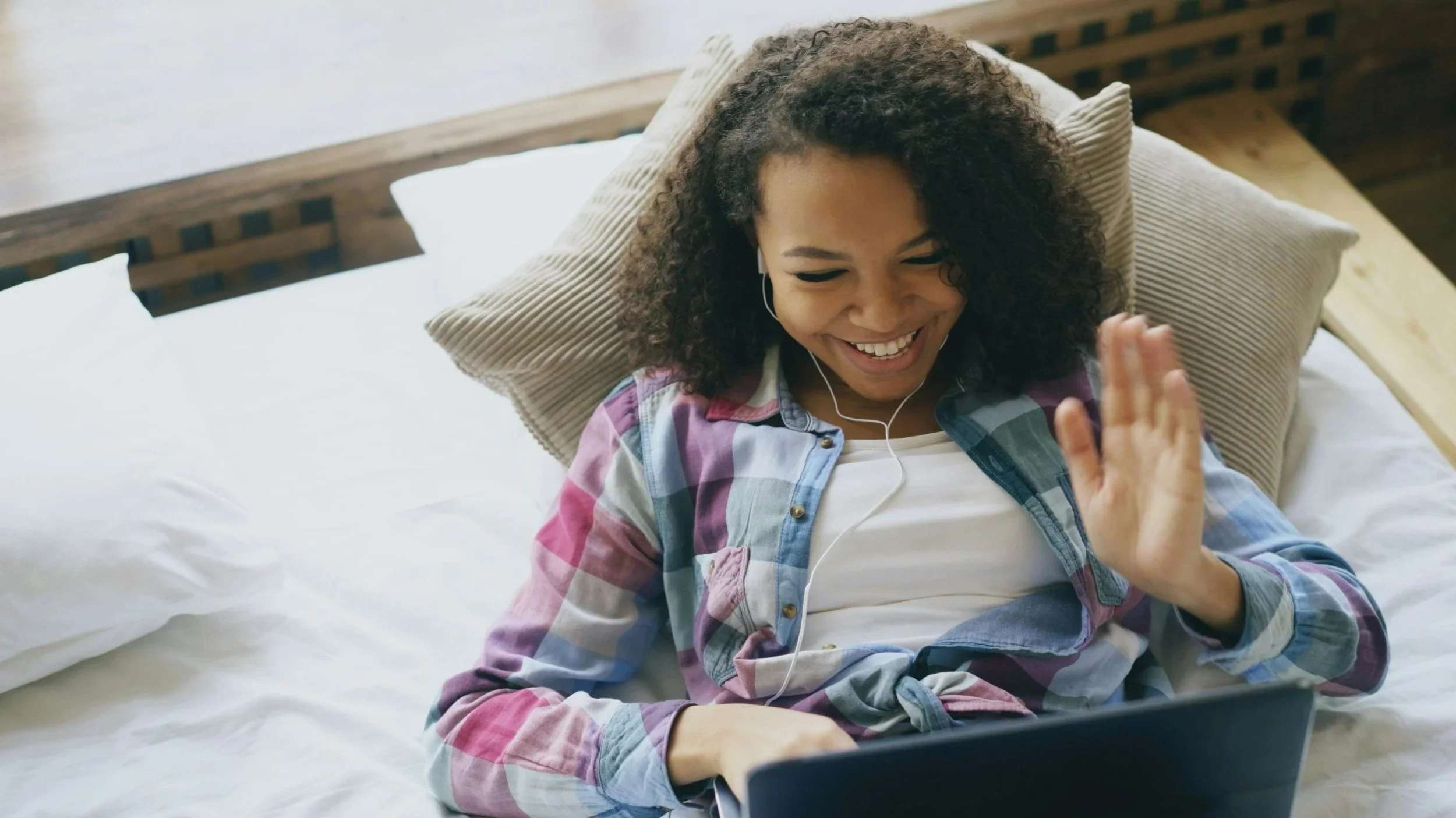 Smiling woman wearing earbuds and waving during a video call on a laptop in bed