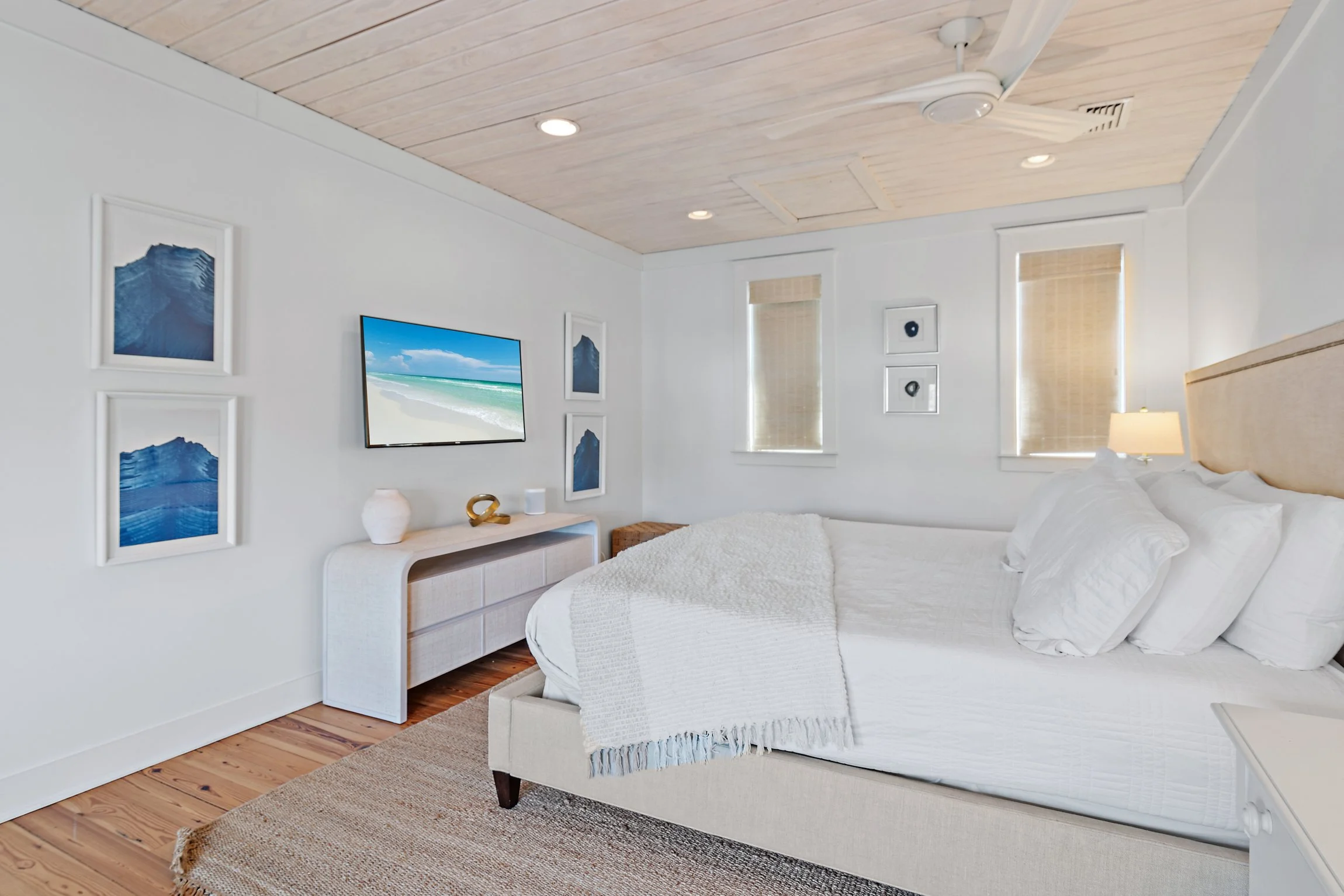 Elegant coastal bedroom featuring a plush king-size bed, white linens, natural wood accents, and ocean-inspired artwork. A mounted TV displays a serene beach view, enhancing the tranquil atmosphere.