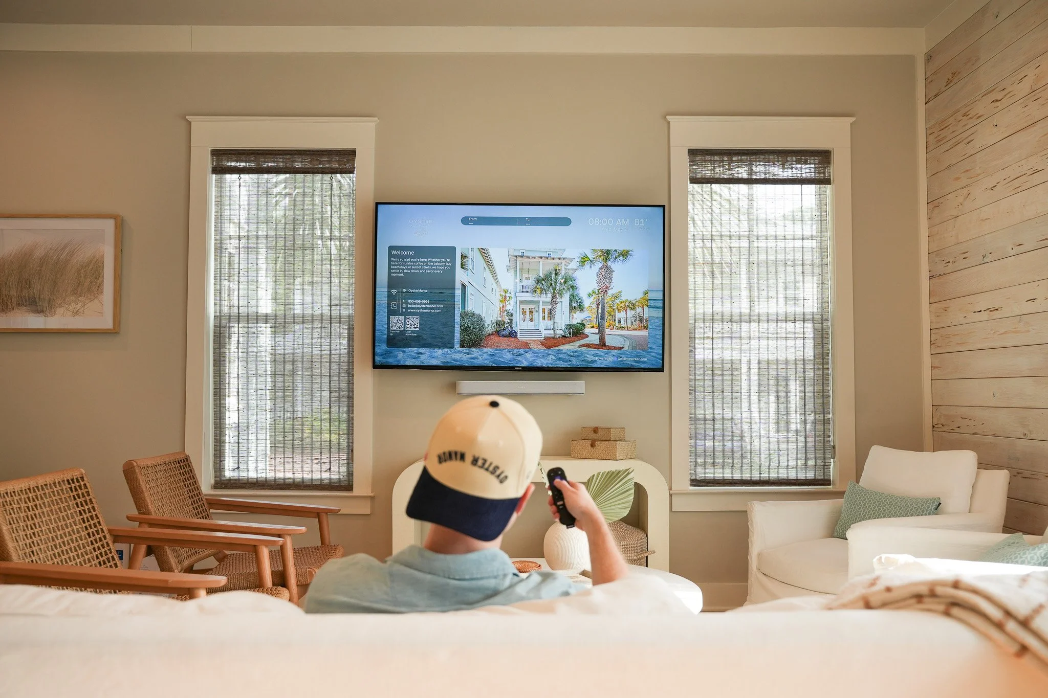 A guest in a casual cap relaxes on a cream sofa while viewing the Oyster Manor Welcome Screen on a mounted living room TV.