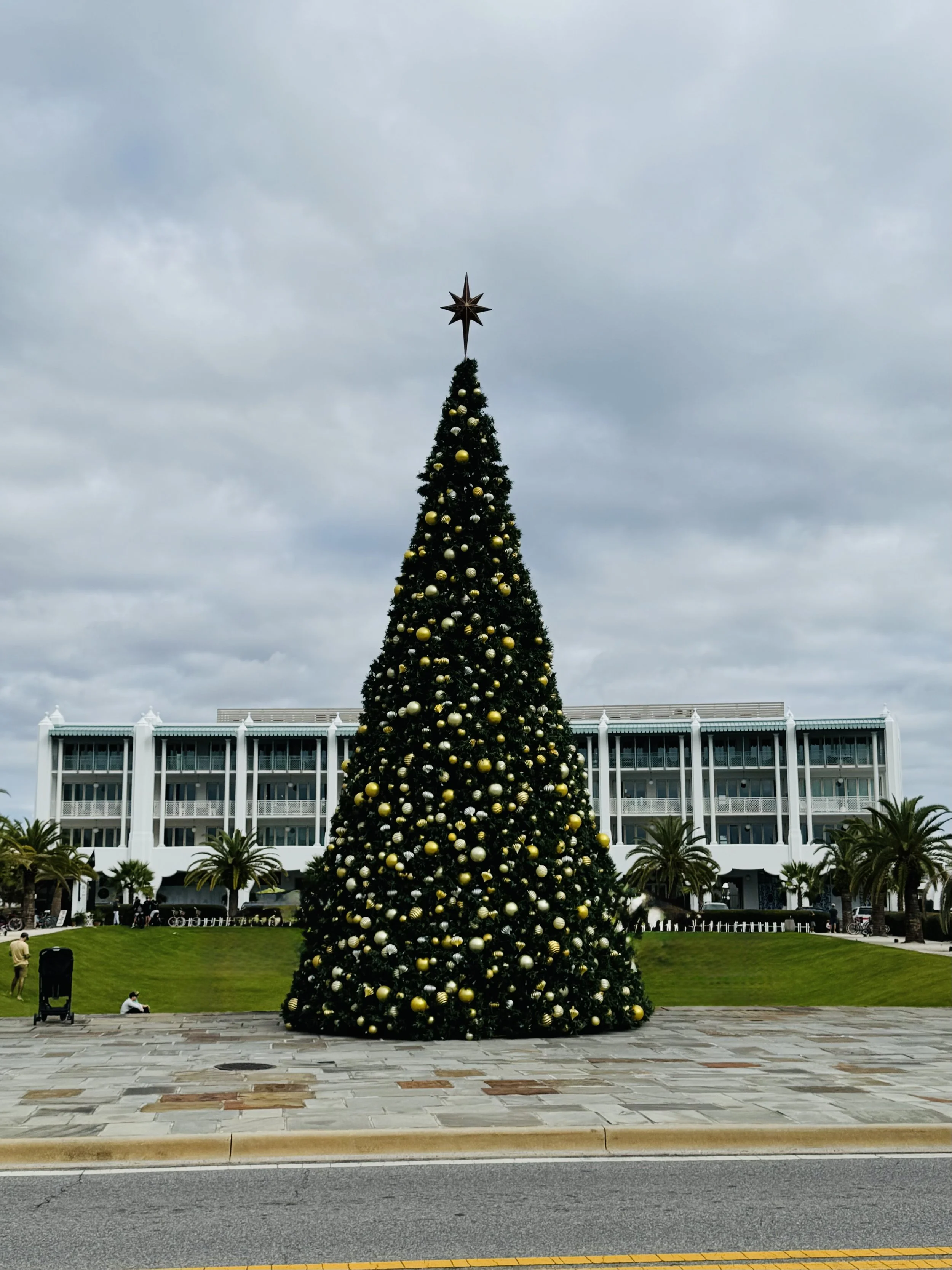 Large decorated Christmas tree at Alys Beach town center with a star topper in front of a white building, surrounded by grass and palm trees.
