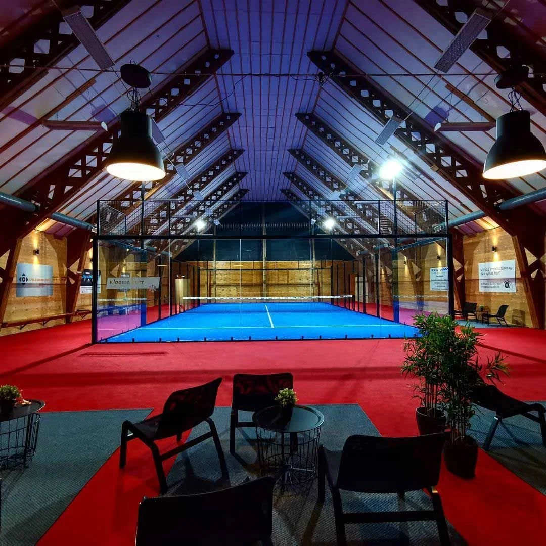 Indoor tennis court with a blue playing surface, enclosed by glass walls, inside a wooden-paneled structure with a sloped ceiling and hanging lights.
