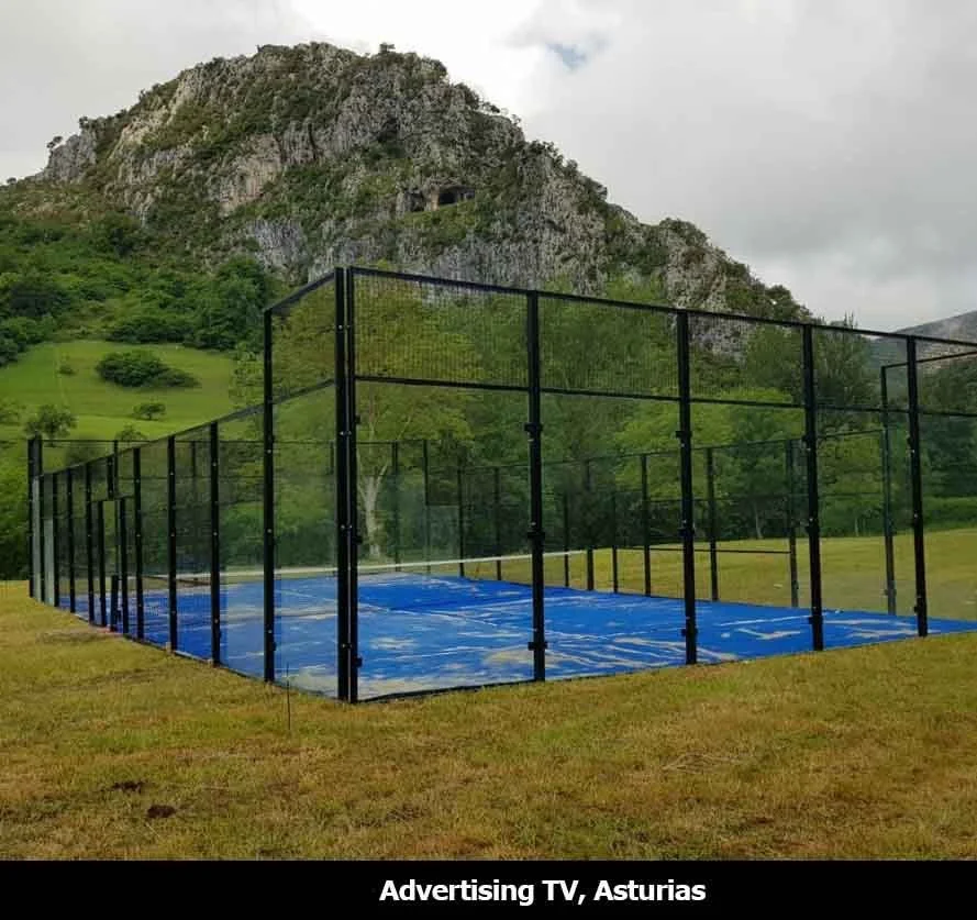 A fenced padel tennis court with a blue surface set in a grassy area, with a green mountain and cloudy sky in the background, in Asturias.