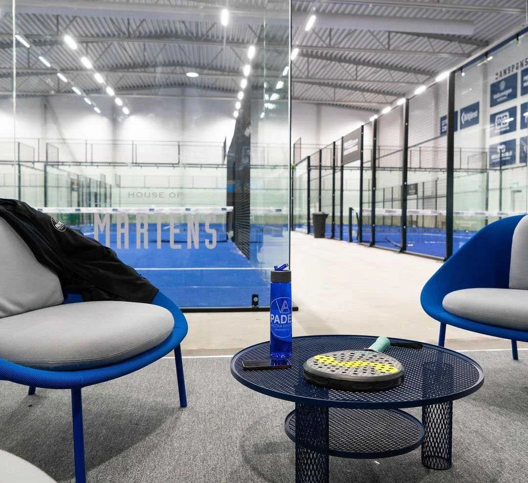 Indoor sports facility with a padel tennis court enclosed by glass walls, and viewing area with two blue and gray chairs, a black metal table holding a blue water bottle, a racket, a shuttlecock, and a small towel.