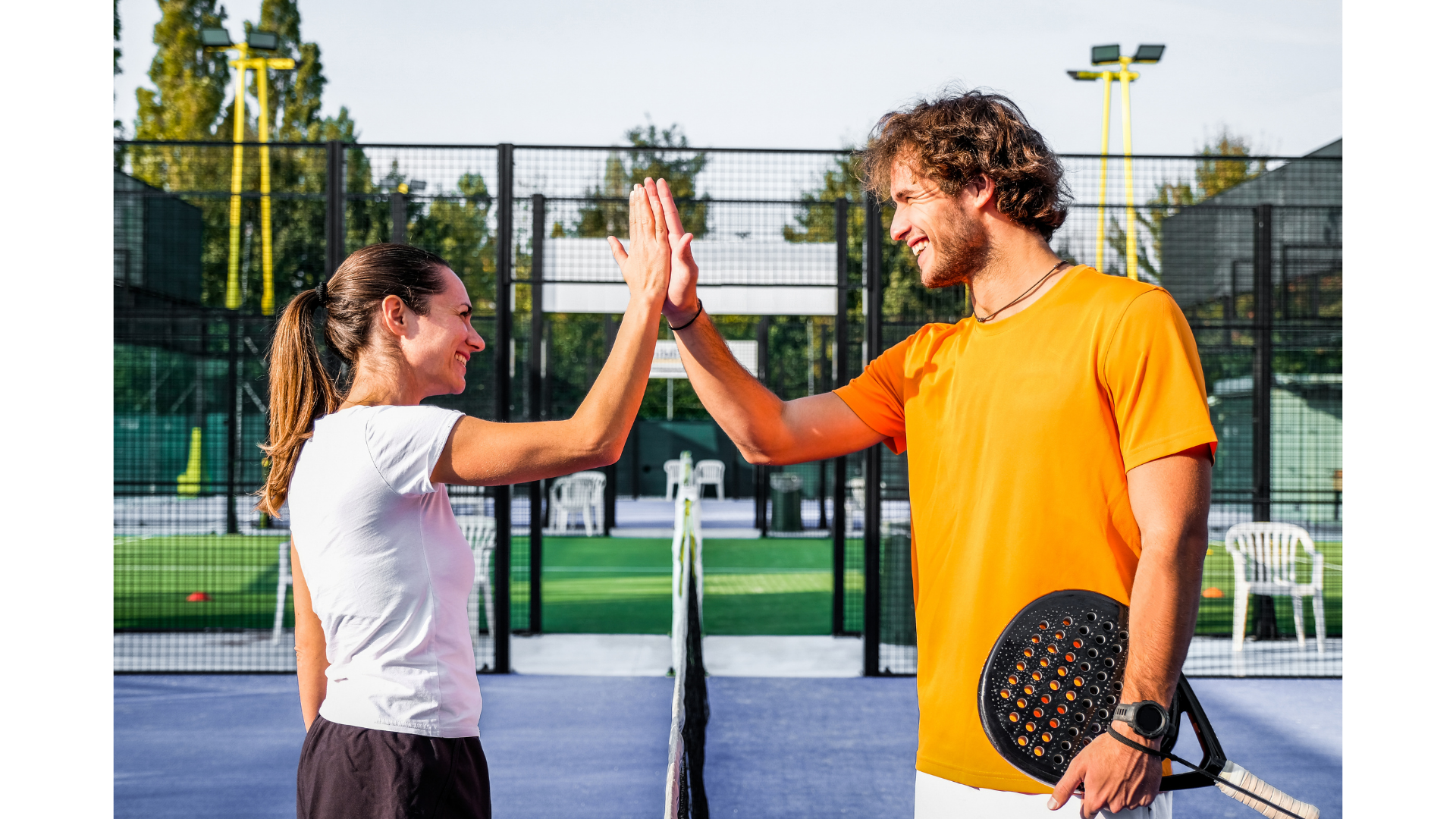 A white man and woman stand high fiving over a padel court net. The sun is out and both are smiling dressed in sports gear.
