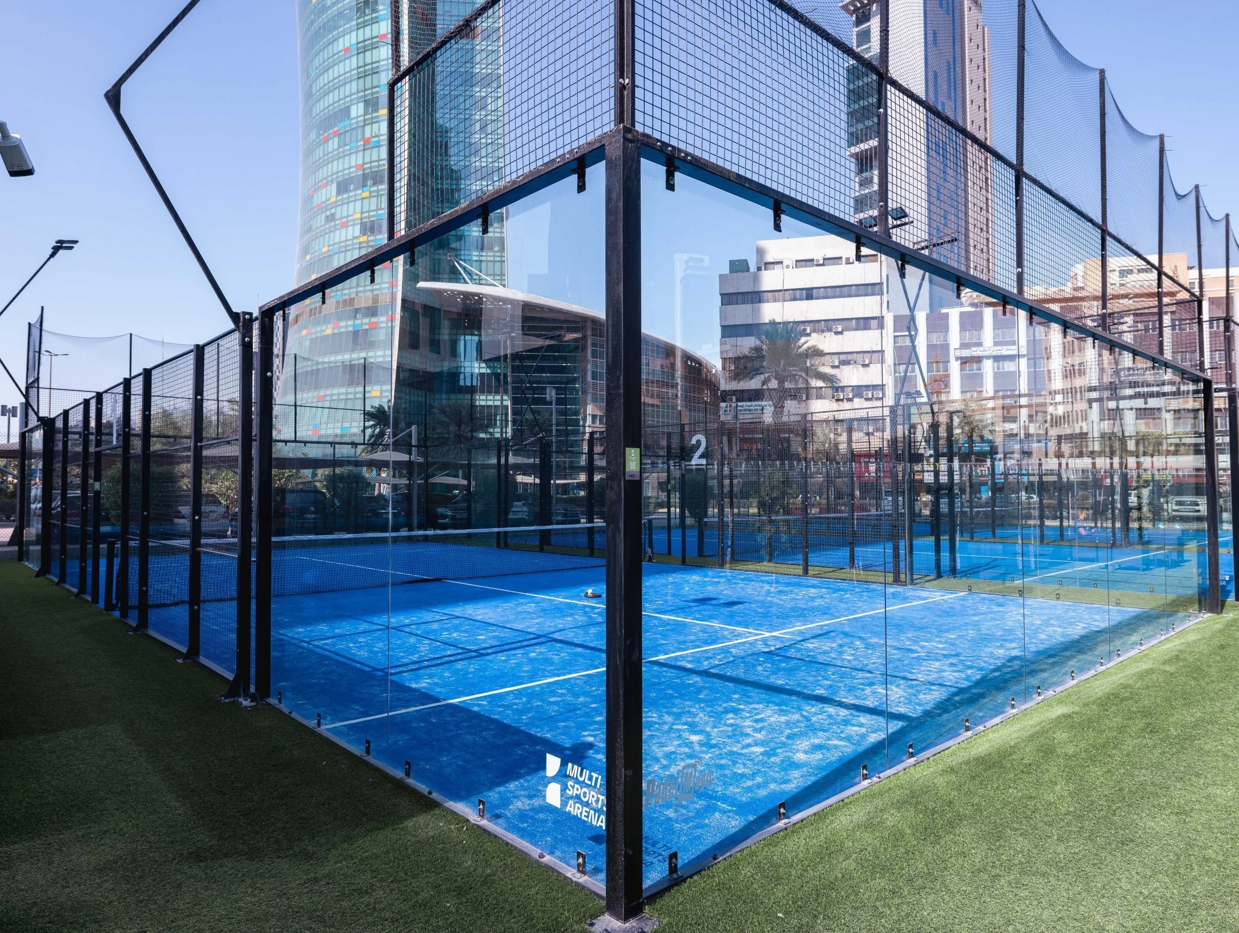 Outdoor blue padel tennis court surrounded by glass panels in an urban setting with tall buildings and city streets in the background.