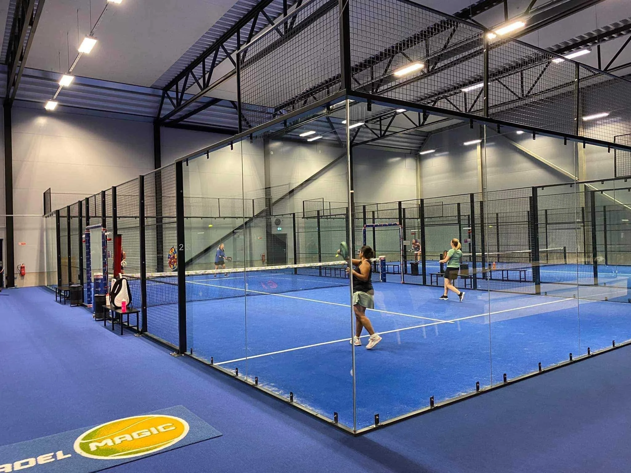 Indoor padel tennis courts with four players practicing, enclosed by glass walls, with seating and equipment along the side.