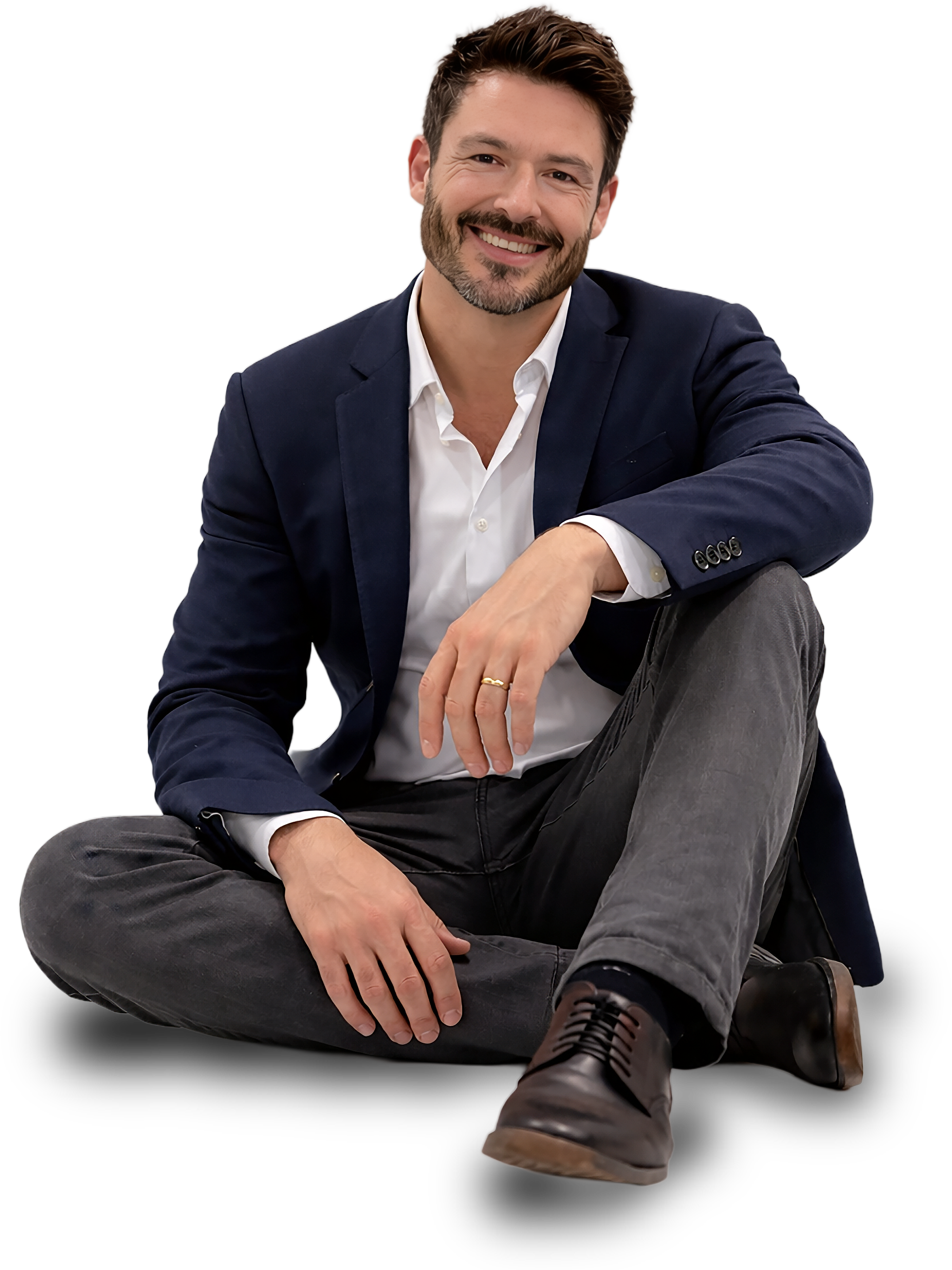 A man with brown hair and a beard sits cross-legged, smiling at the camera. He is dressed in a dark blazer, white shirt, gray pants, and dark shoes.