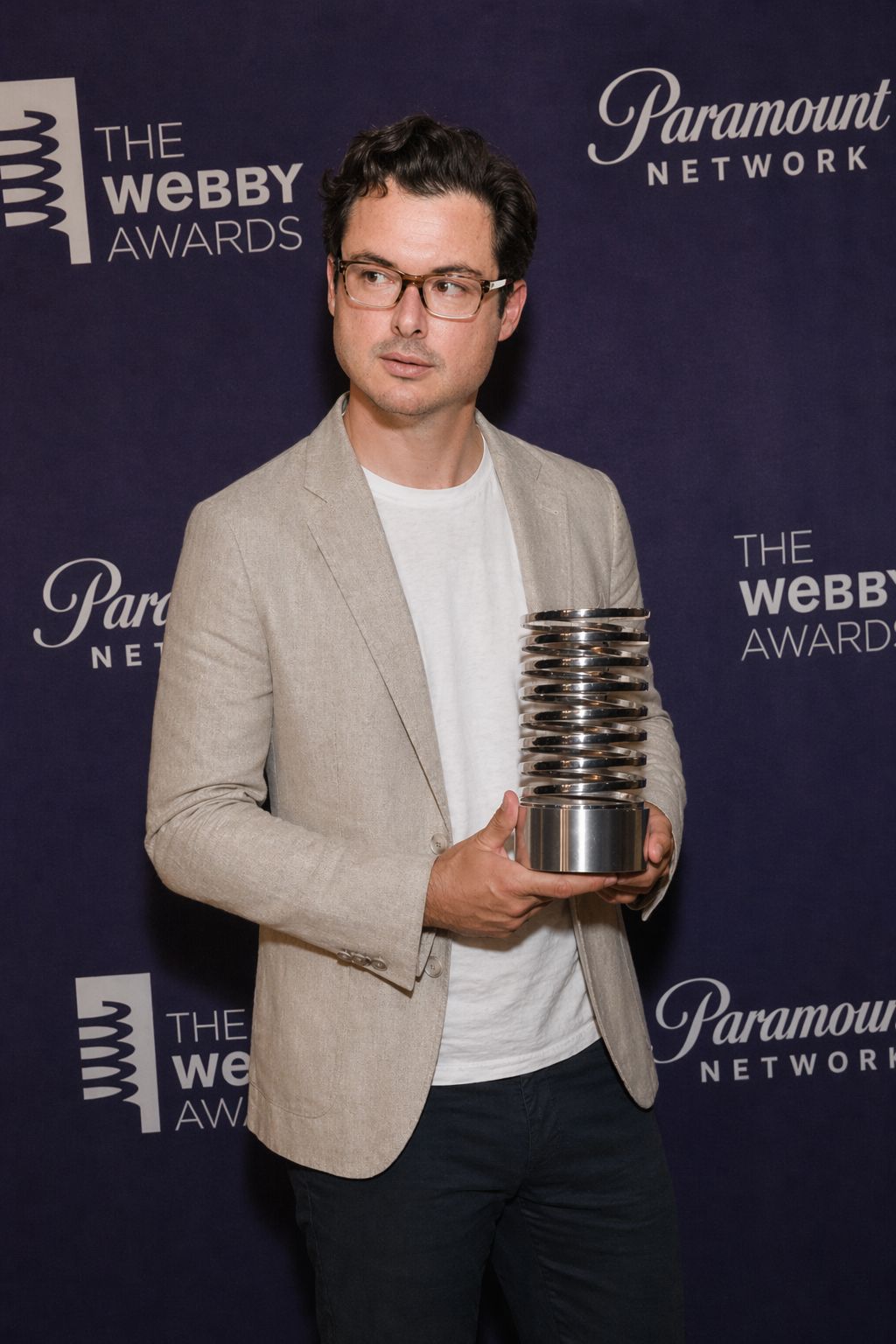 A man in glasses and a beige blazer holding a silver Webby Award trophy at the Webby Awards event.