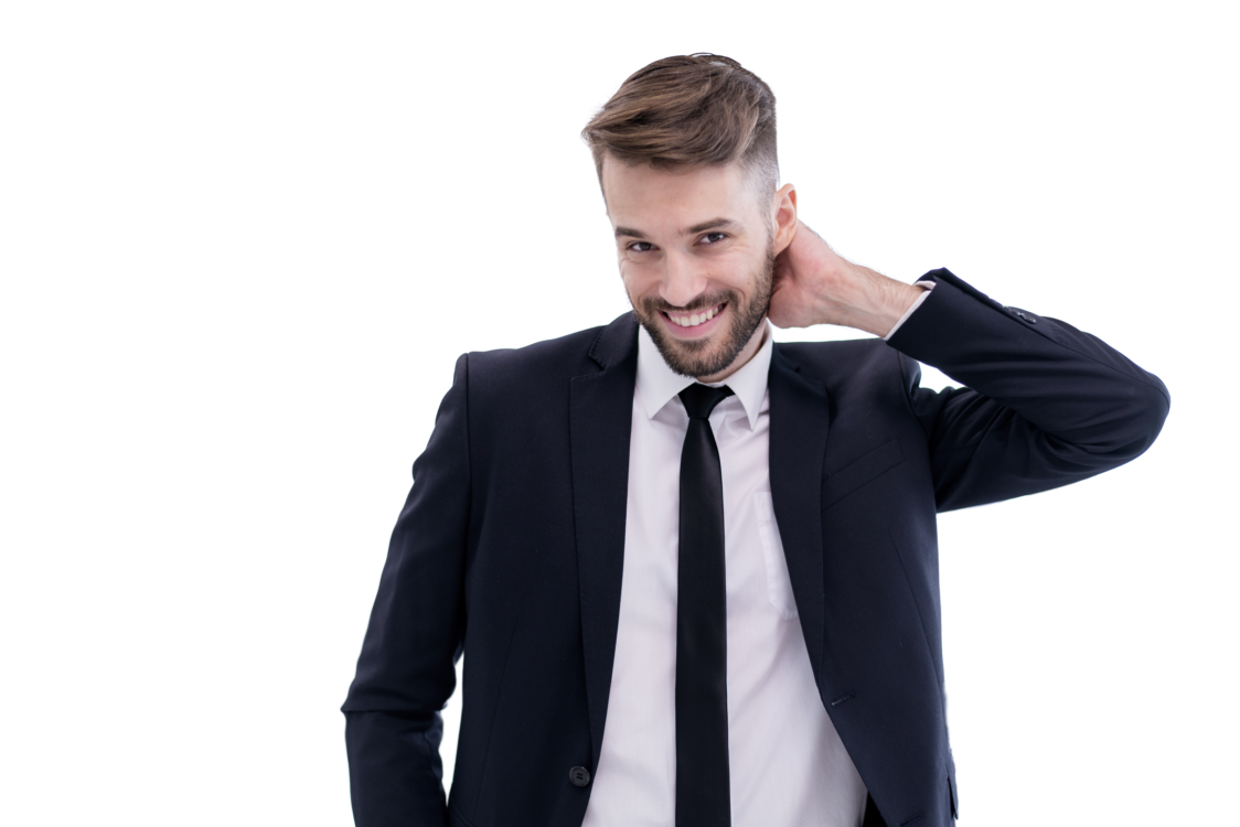 A young man in a black suit, white shirt, and black tie smiling and touching the back of his neck.