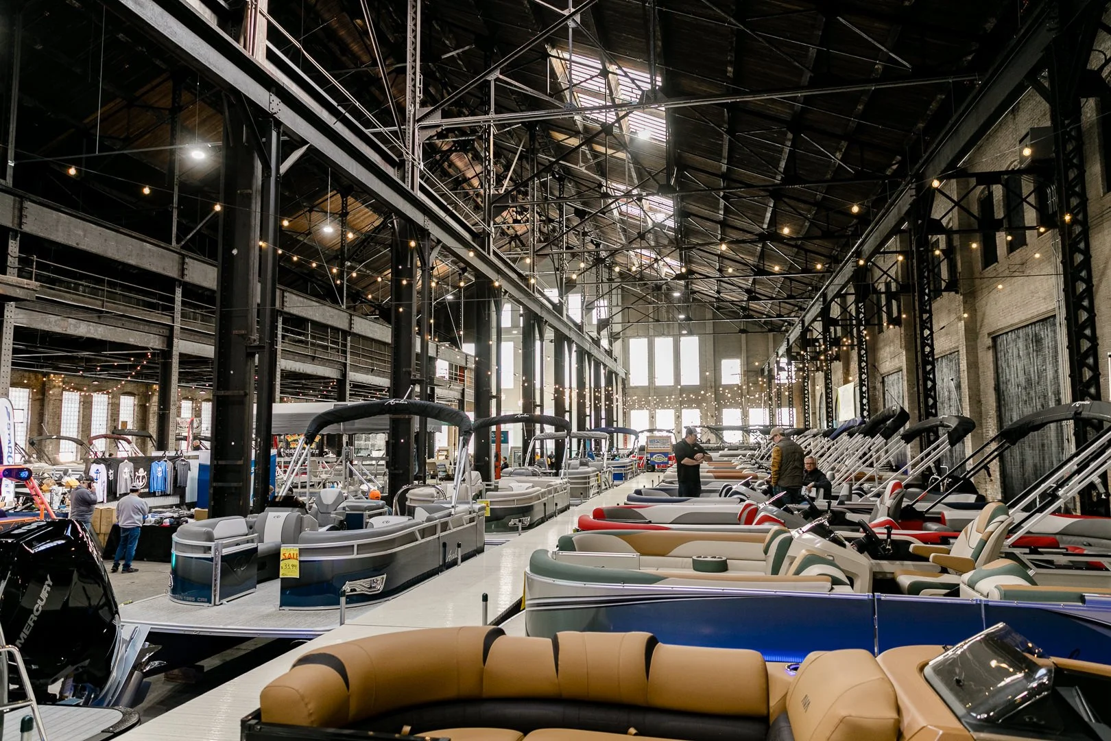 Indoor boat show with various boats, RVs, and people walking around in a large industrial-style building with high ceilings, exposed beams, and large windows.