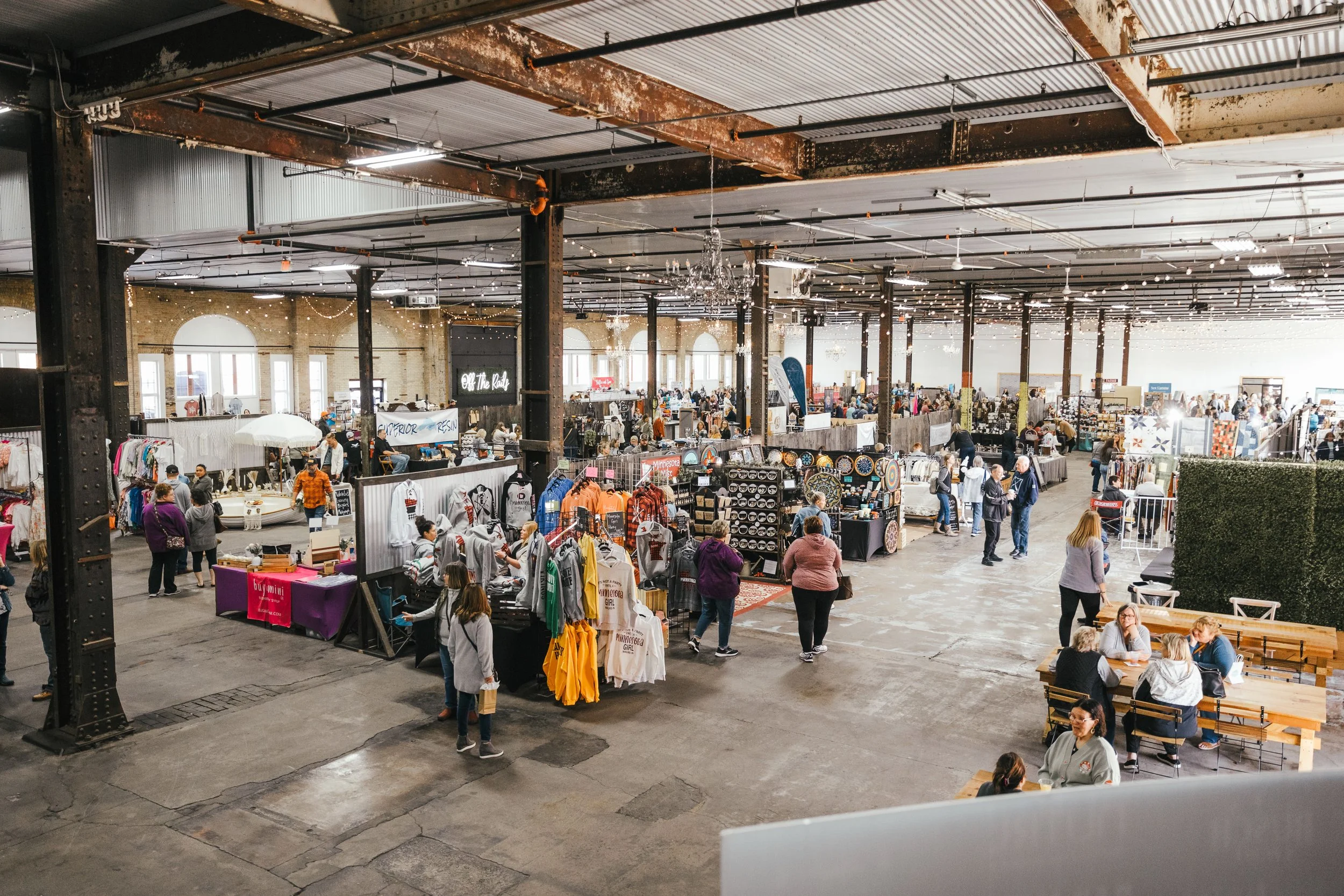 Interior of a large indoor market with various booths selling clothing, accessories, and crafts. People are browsing, shopping, and sitting at tables. The space has exposed beams, industrial decor, and string lights.