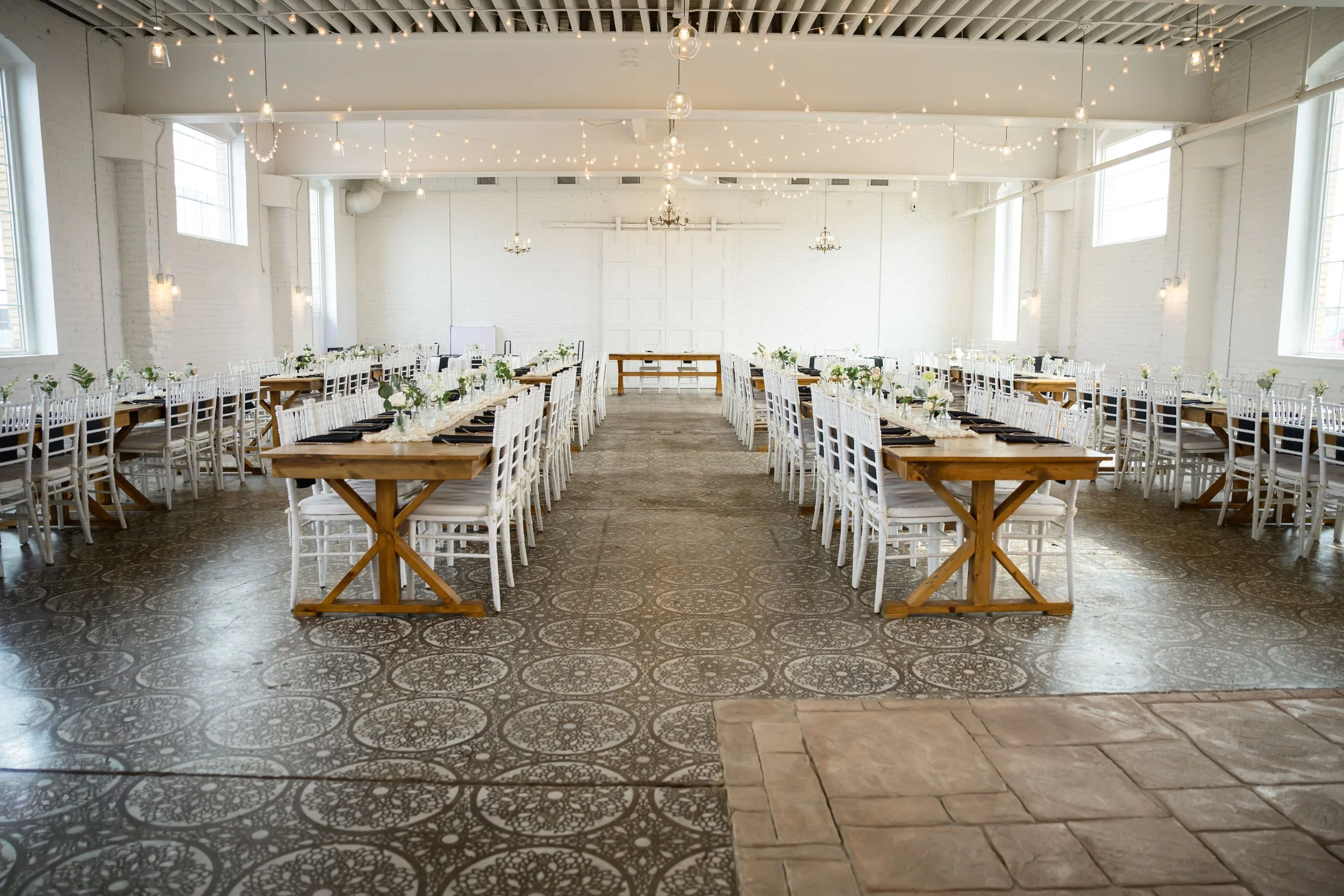 Empty wedding reception hall with rows of light wood chairs, decorated tables with floral centerpieces, white brick walls, large windows, and string lights hanging from the ceiling. Only available through 2026.
