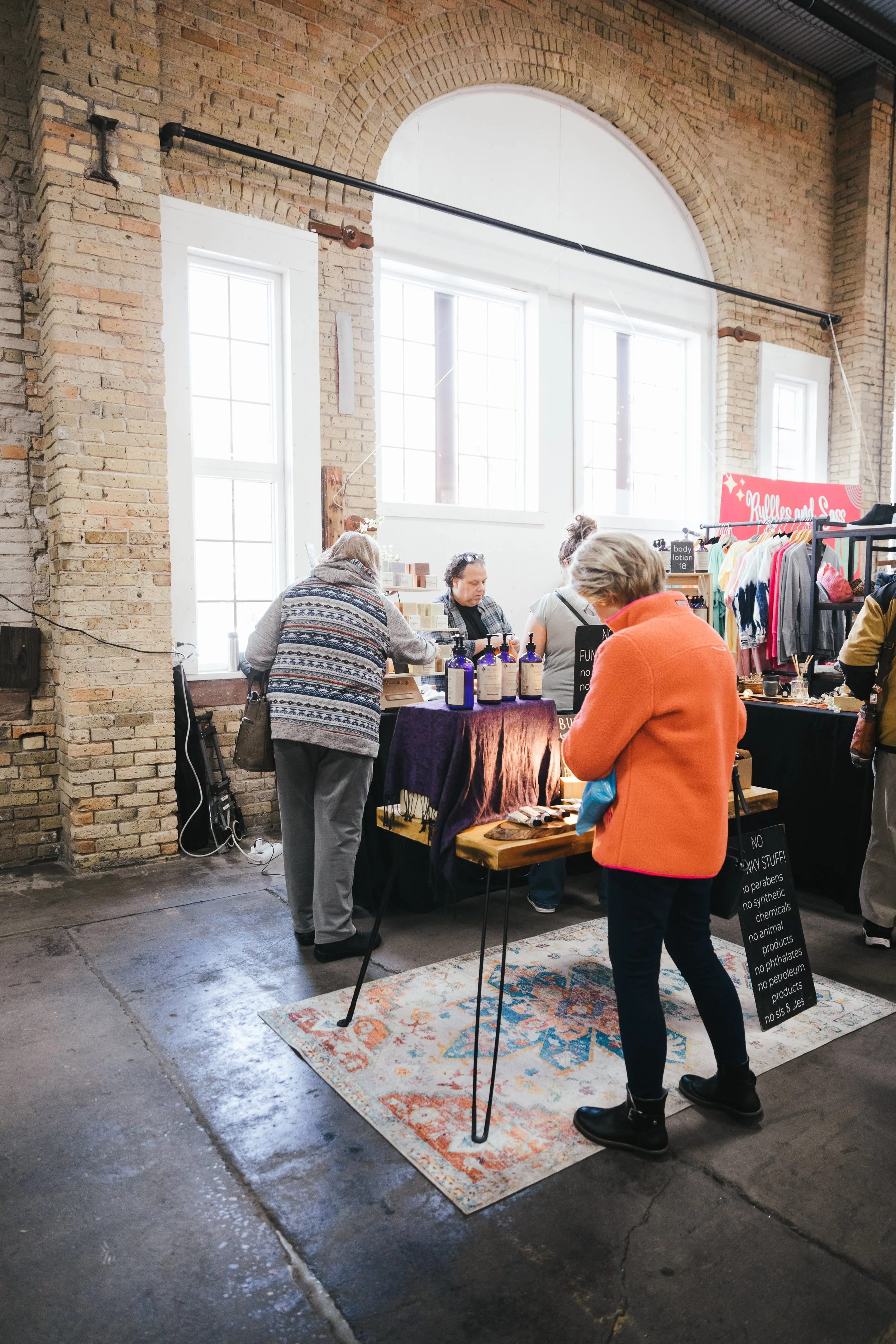People shopping at a booth inside a store with large windows, exposed brick walls, and a colorful rug on concrete floor.