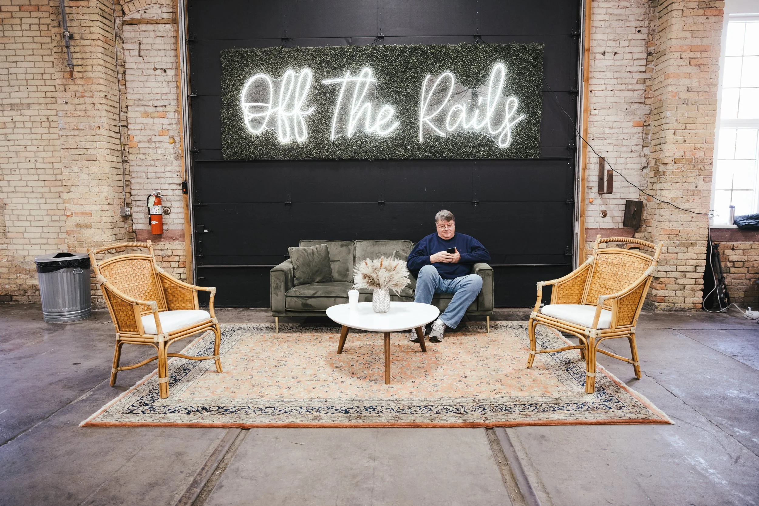 A man sitting on a green couch in a spacious industrial-style room, looking at his phone. Behind him, a neon sign reads 'Off The Rails' on a textured wall. In front of the couch is a round white coffee table with a vase of dried flowers. Two wicker c