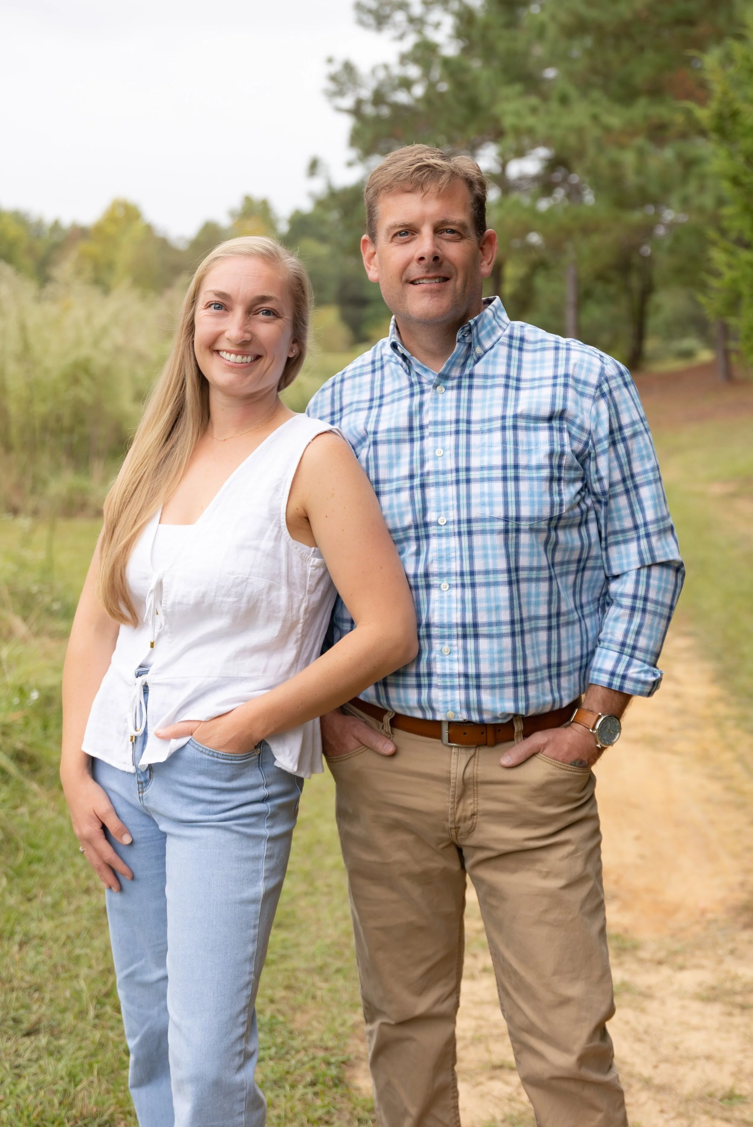 A smiling couple standing on a dirt path in a green park or forest during daytime.