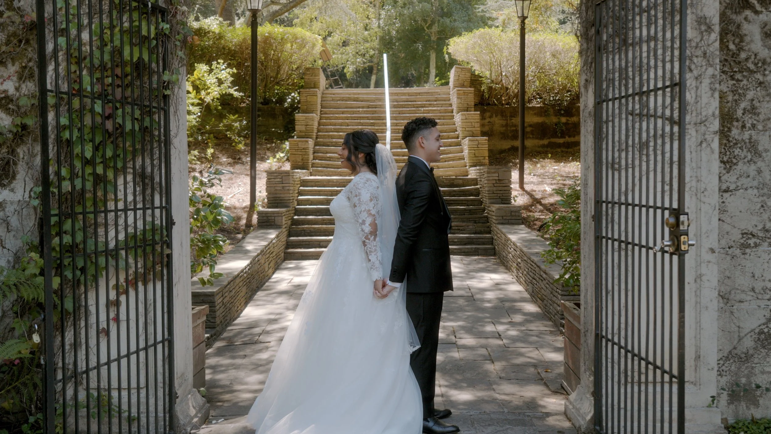 A bride and groom stand back to back, holding hands, in a garden with stairs and greenery in the background.