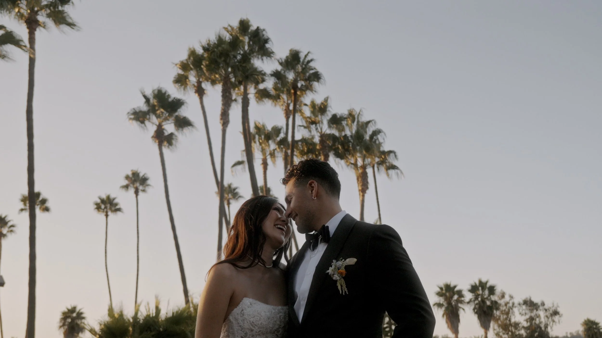 A newlywed couple in wedding attire sharing a tender moment outdoors during sunset, with tall palm trees in the background.