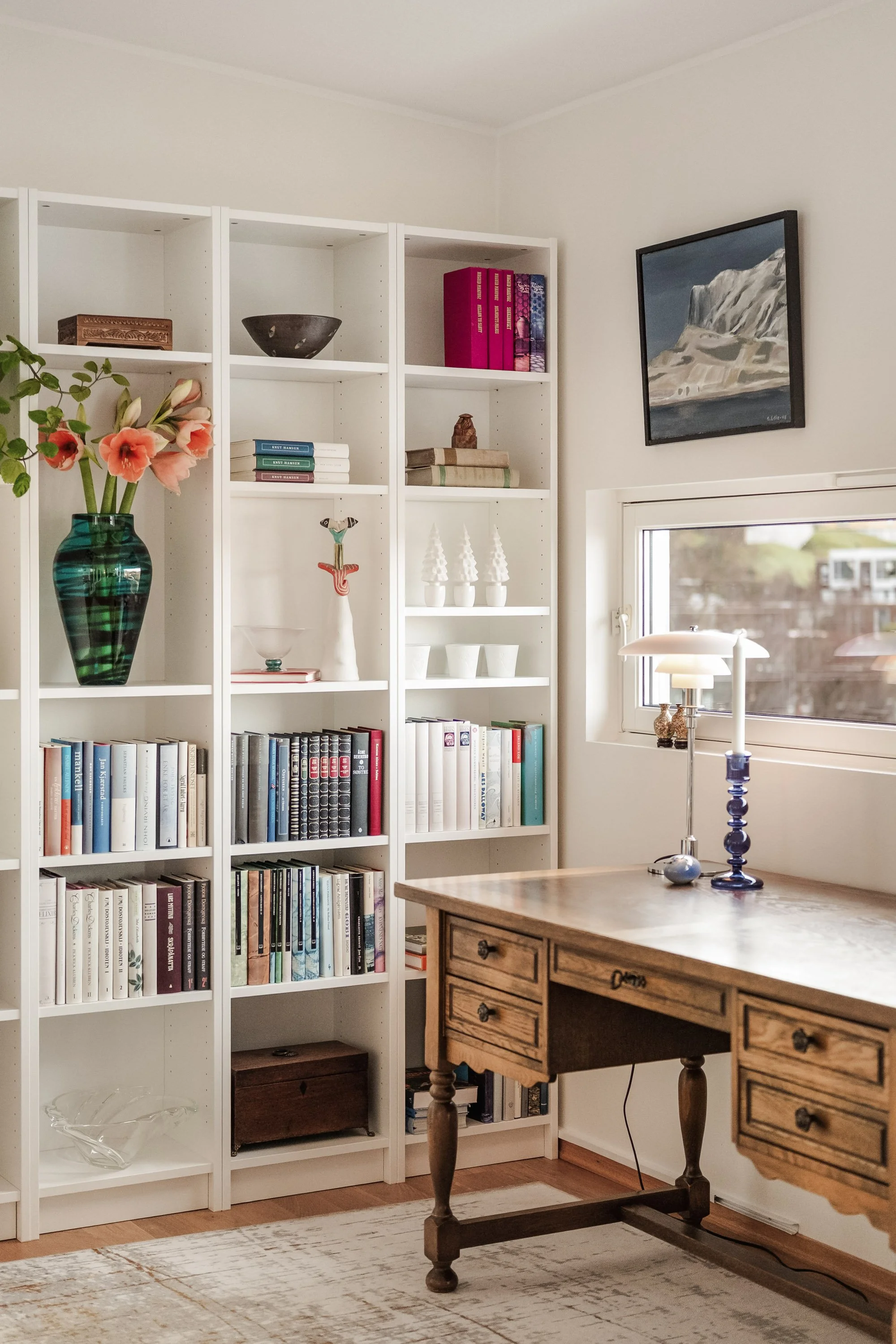 Et innendørsmiljø med bøker og dekorative gjenstander på hvite hyller, et skrivebord og et vindu.