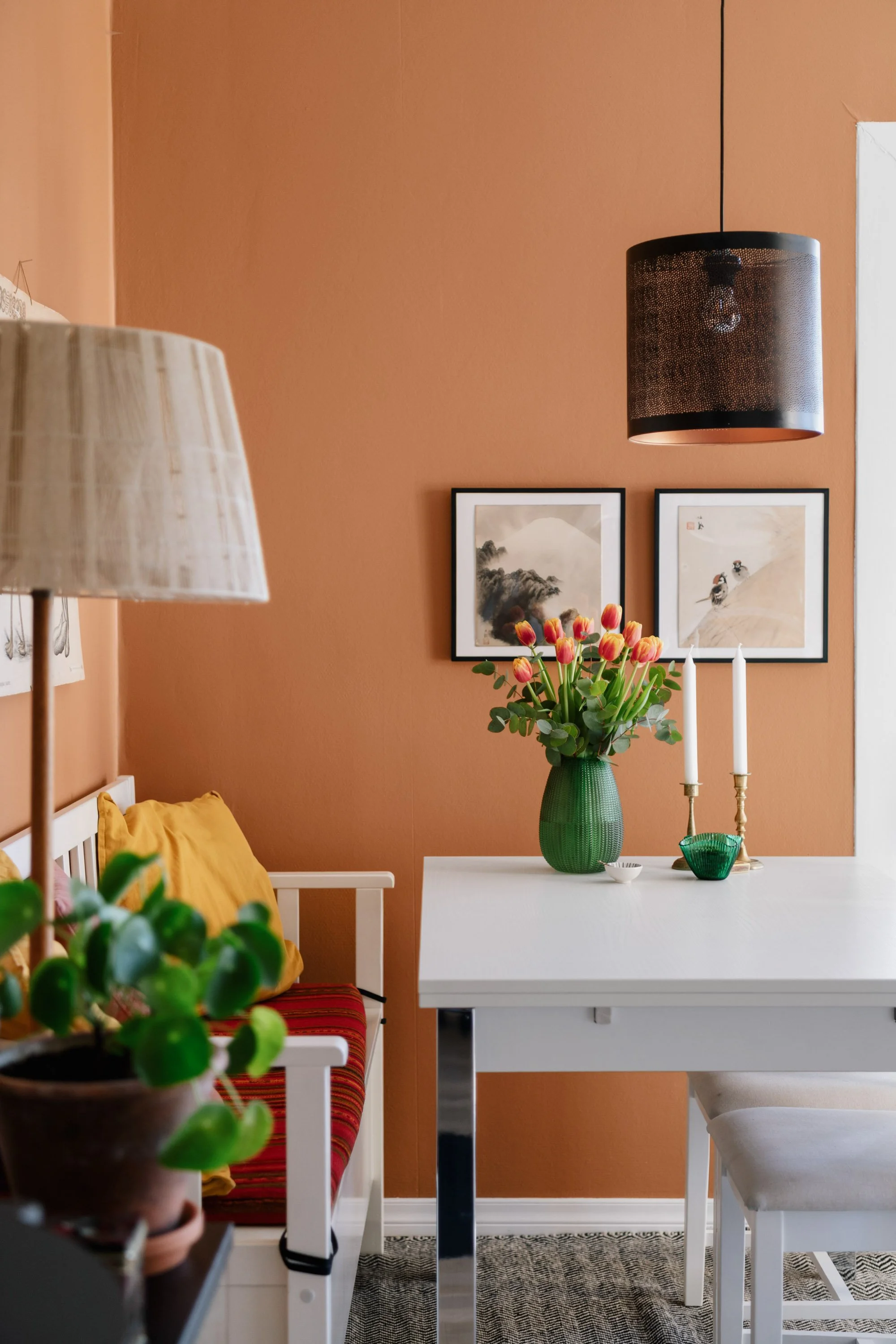 Et spisebord med en grønn vase med roser, to lysestaker, og illustrasjonsbilder på en terrakottafarget vegg.