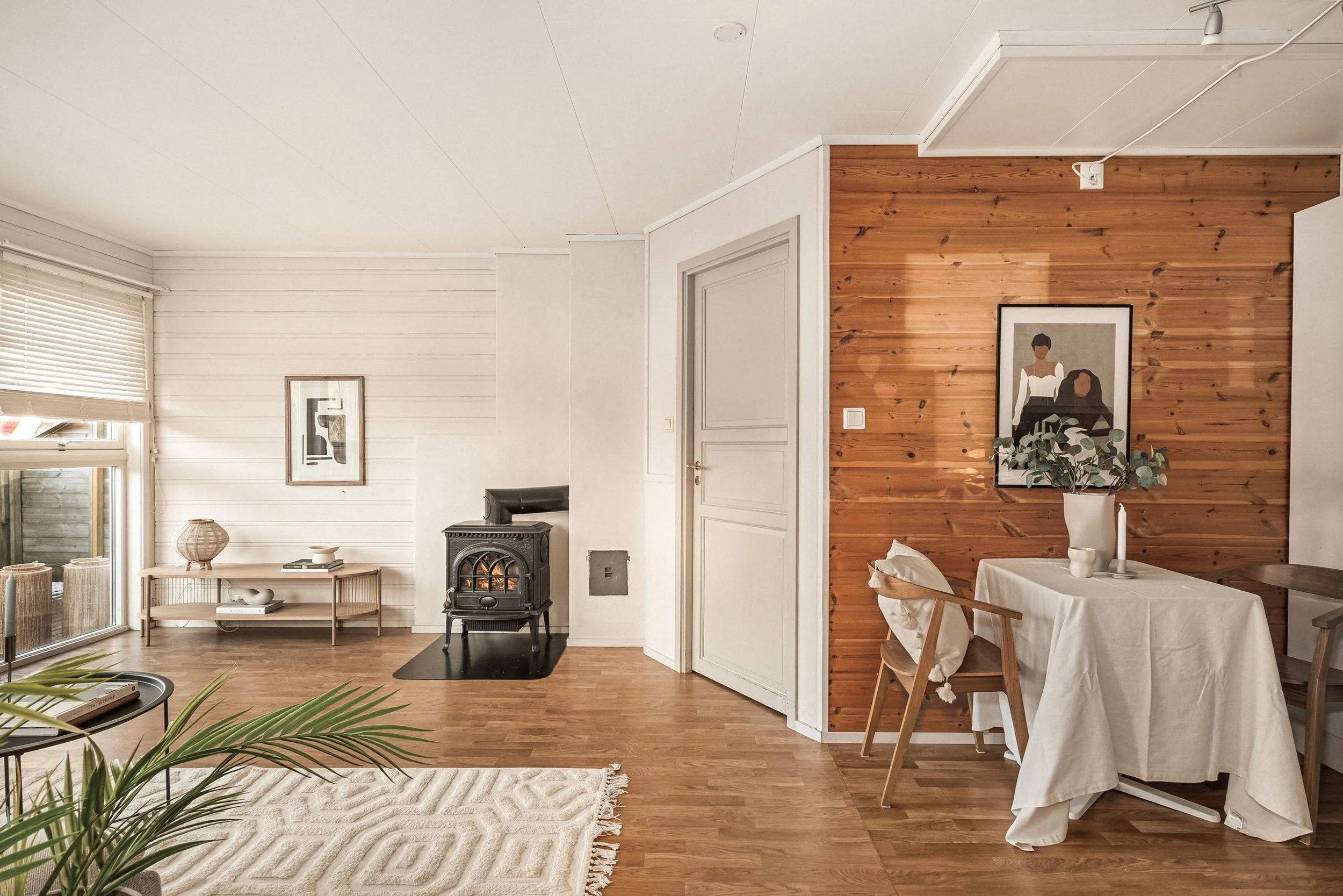 Lys stue med hvite vegger, tregulv, peis, spisebord med hvitt laken, vaser og kunst på veggen.