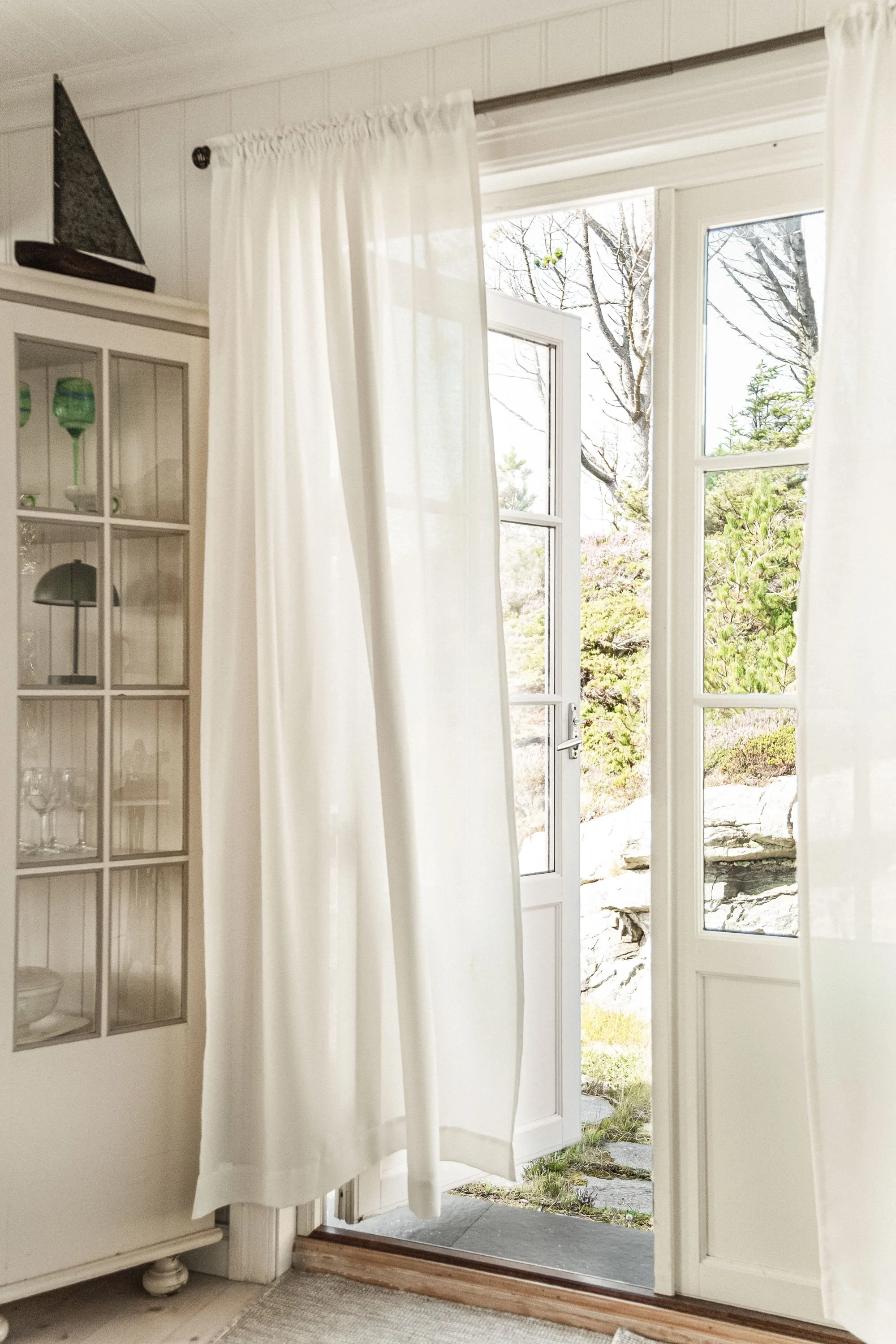 Et hvitt rom med vinduer og en åpen dør som fører ut til en hage med stein og grønne planter, samt en hvit skyvedørsgardin.