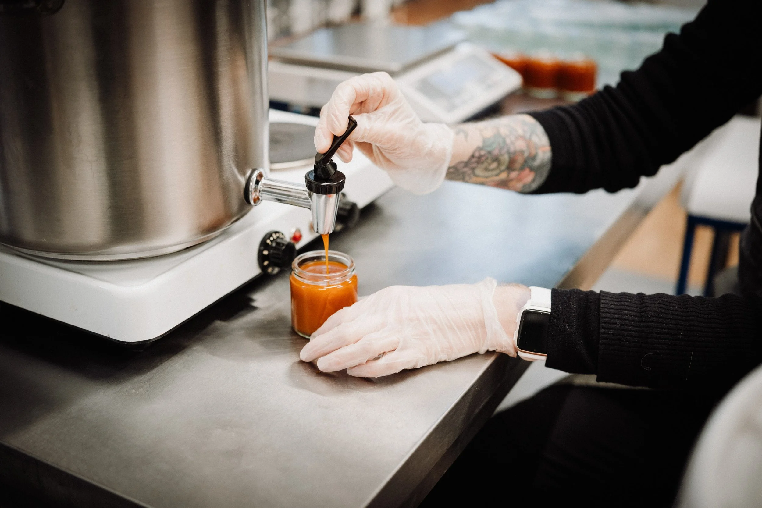 En person overfører en oransje saus fra en stor metallkar til et lite glasskrukke. Personen har på hvite engangshansker og tatoverte armer, og de står på et metallbord.
